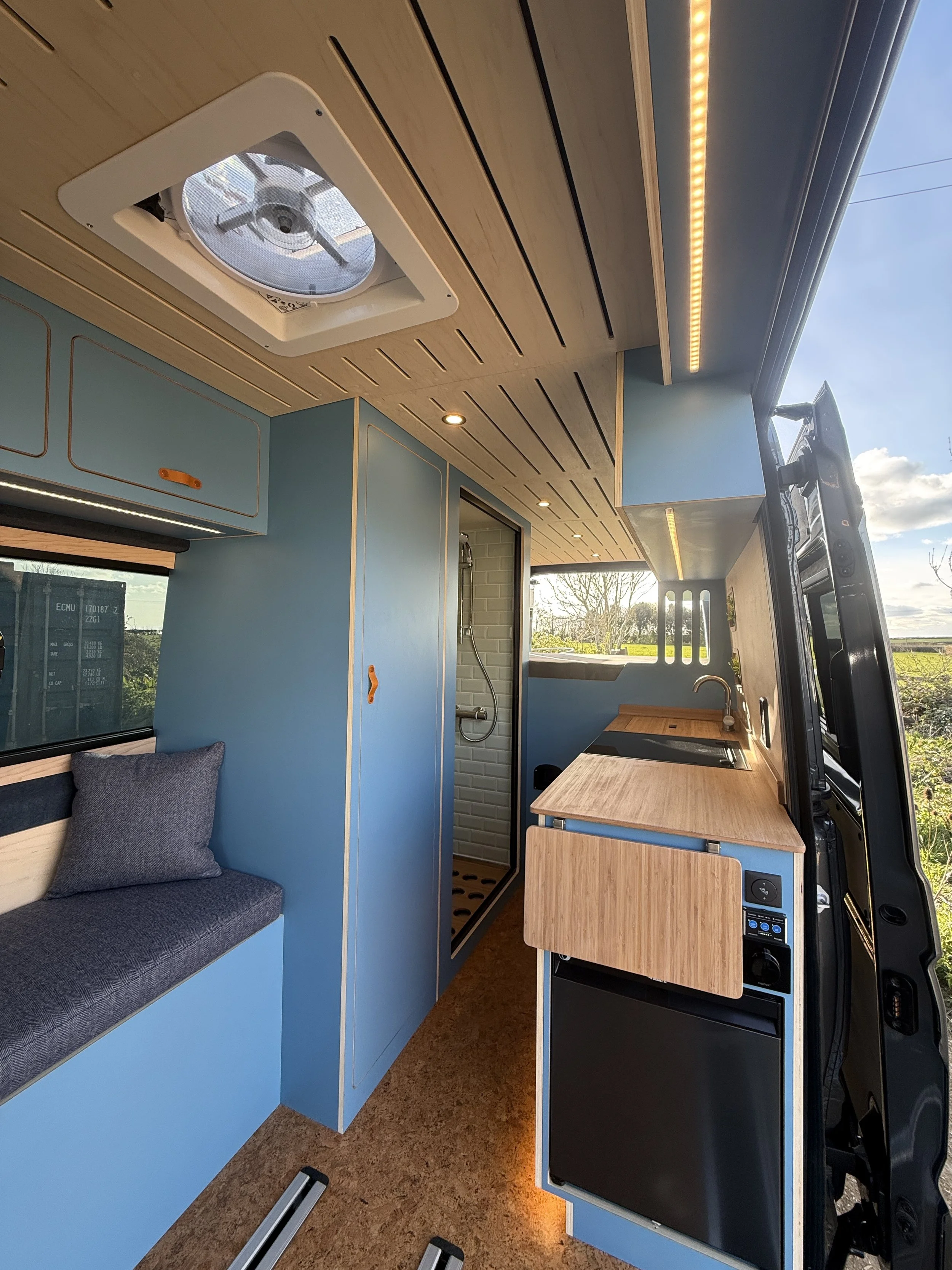 Interior of a converted camper van with a wooden ceiling, blue walls, a small seating area with a cushion, a compact kitchen with a sink, a countertop, and a small fridge, and a shower stall with a showerhead visible through a door.