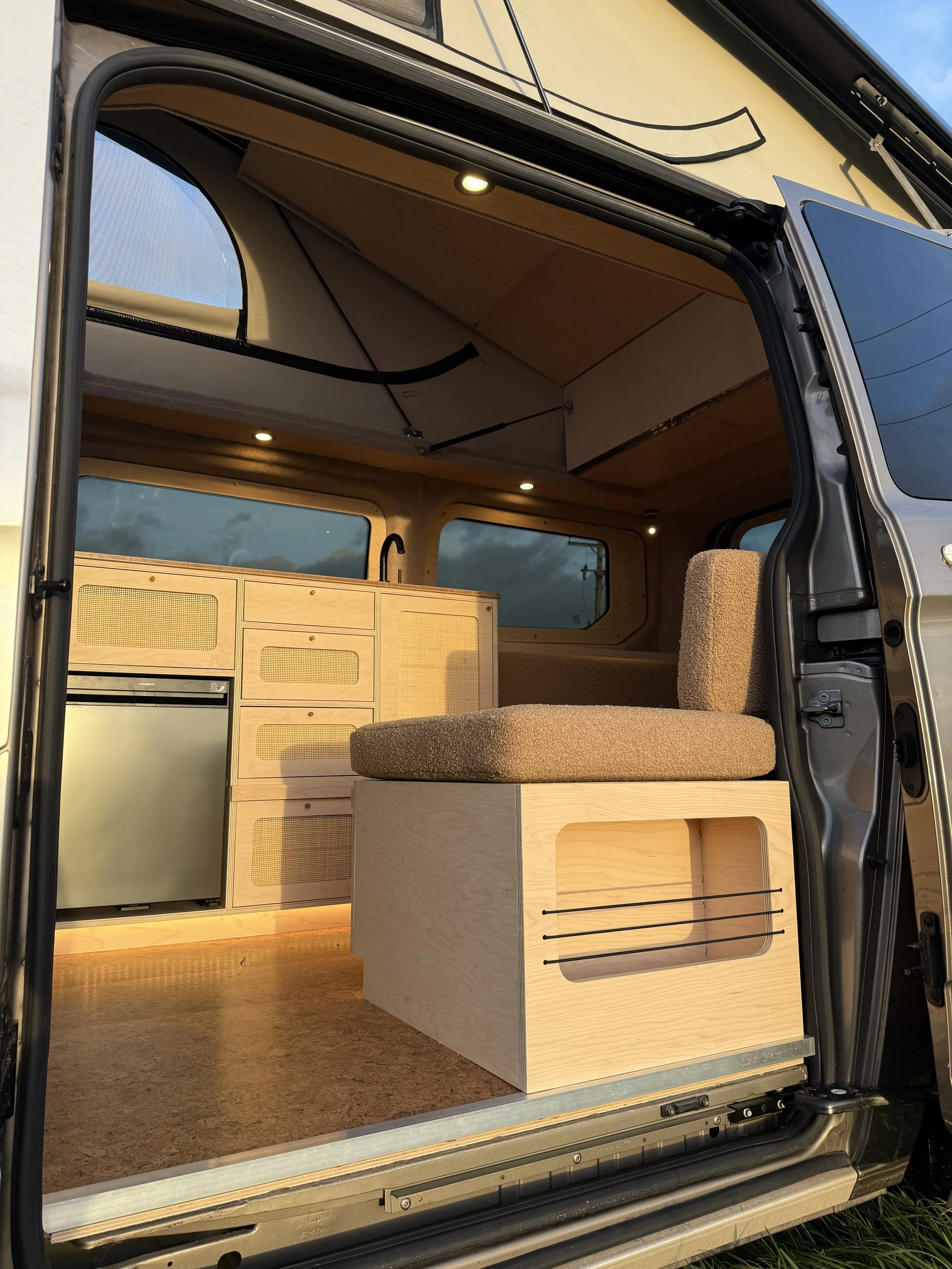 Interior of a camper van with wooden cabinets, a mini refrigerator, a cushioned seat, and interior lighting.