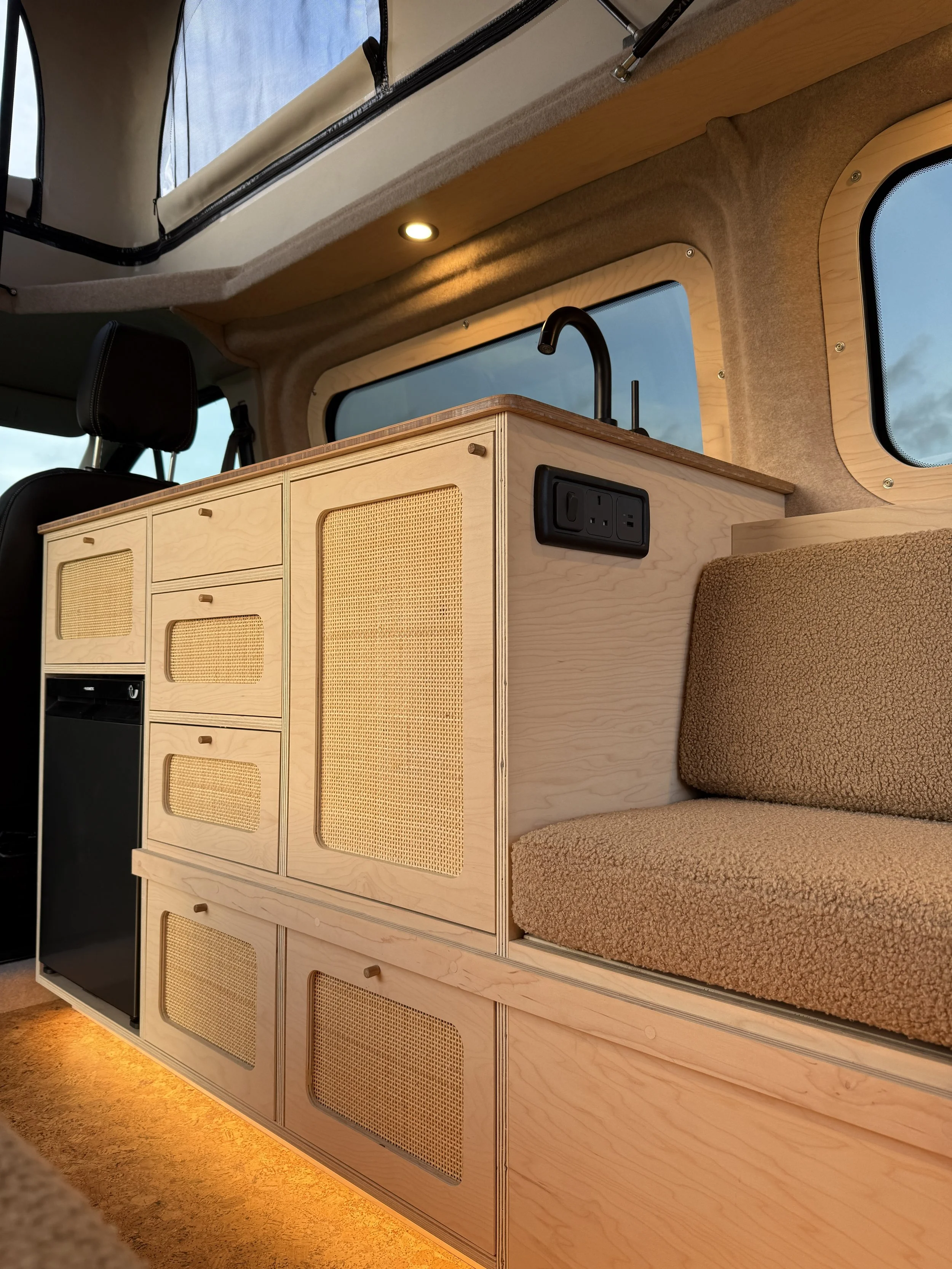 Interior of a camper van showing a wooden cabinet with woven panel doors, a small refrigerator, a seating area with a brown upholstered cushion, and a small sink with a black faucet. There are windows with light-colored wood trim and electrical outle