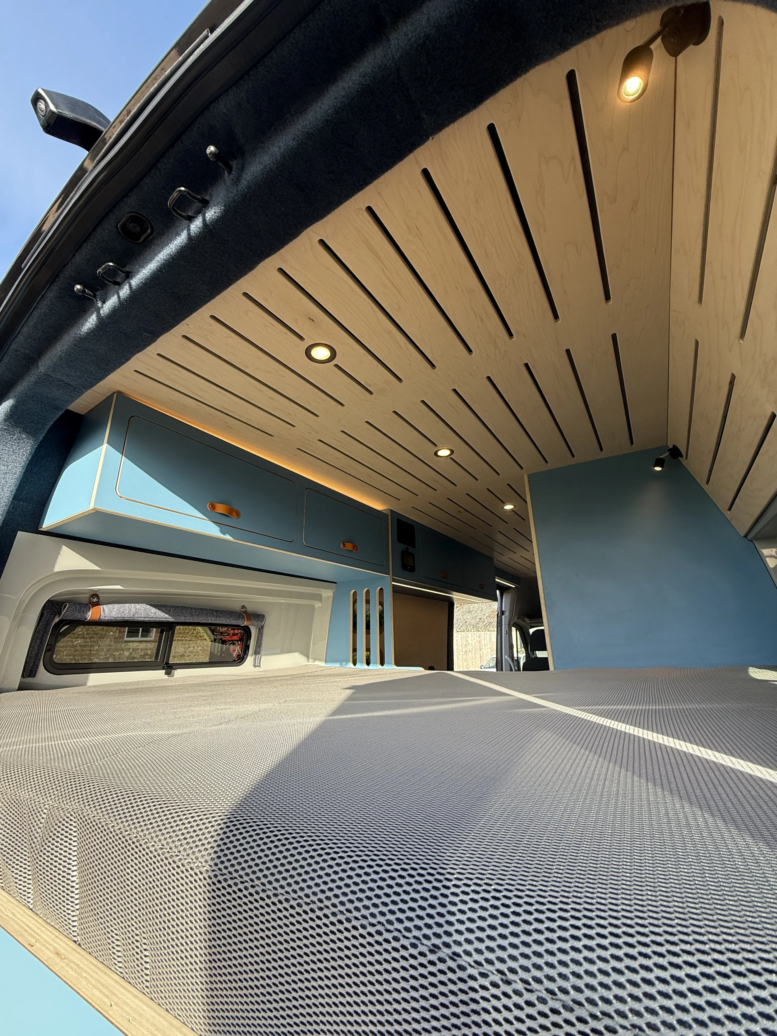 Interior of a camper van with wooden ceiling, blue cabinets, and a bed with a mattress. Small ceiling lights illuminate the space.