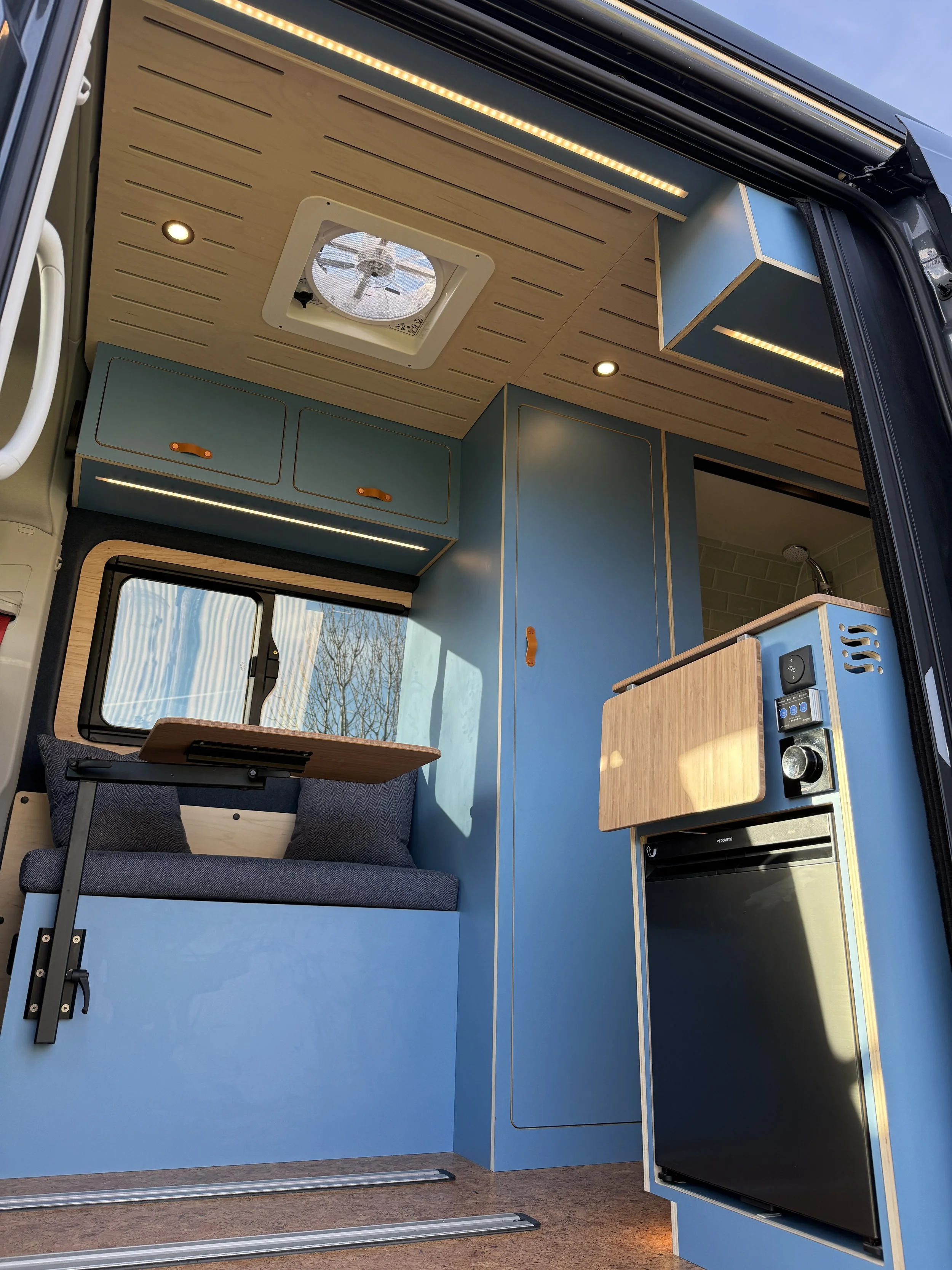Interior view of a compact camper van with blue cabinetry, a small fold-out table, seating area, and kitchen appliances, including a mini refrigerator and a sink, with wooden accents and LED lighting.