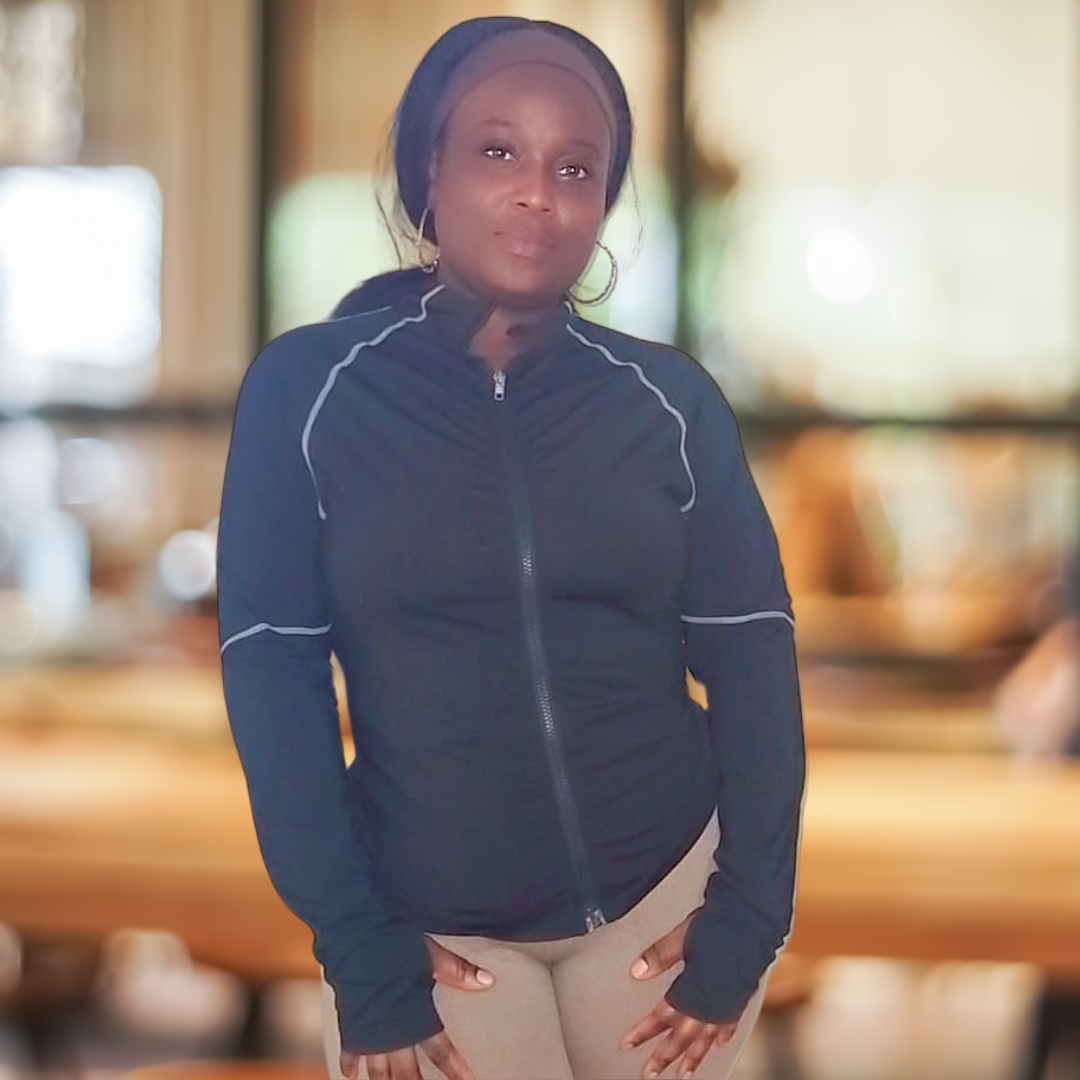 Woman standing indoors wearing a black jacket and beige pants, with blurred background.