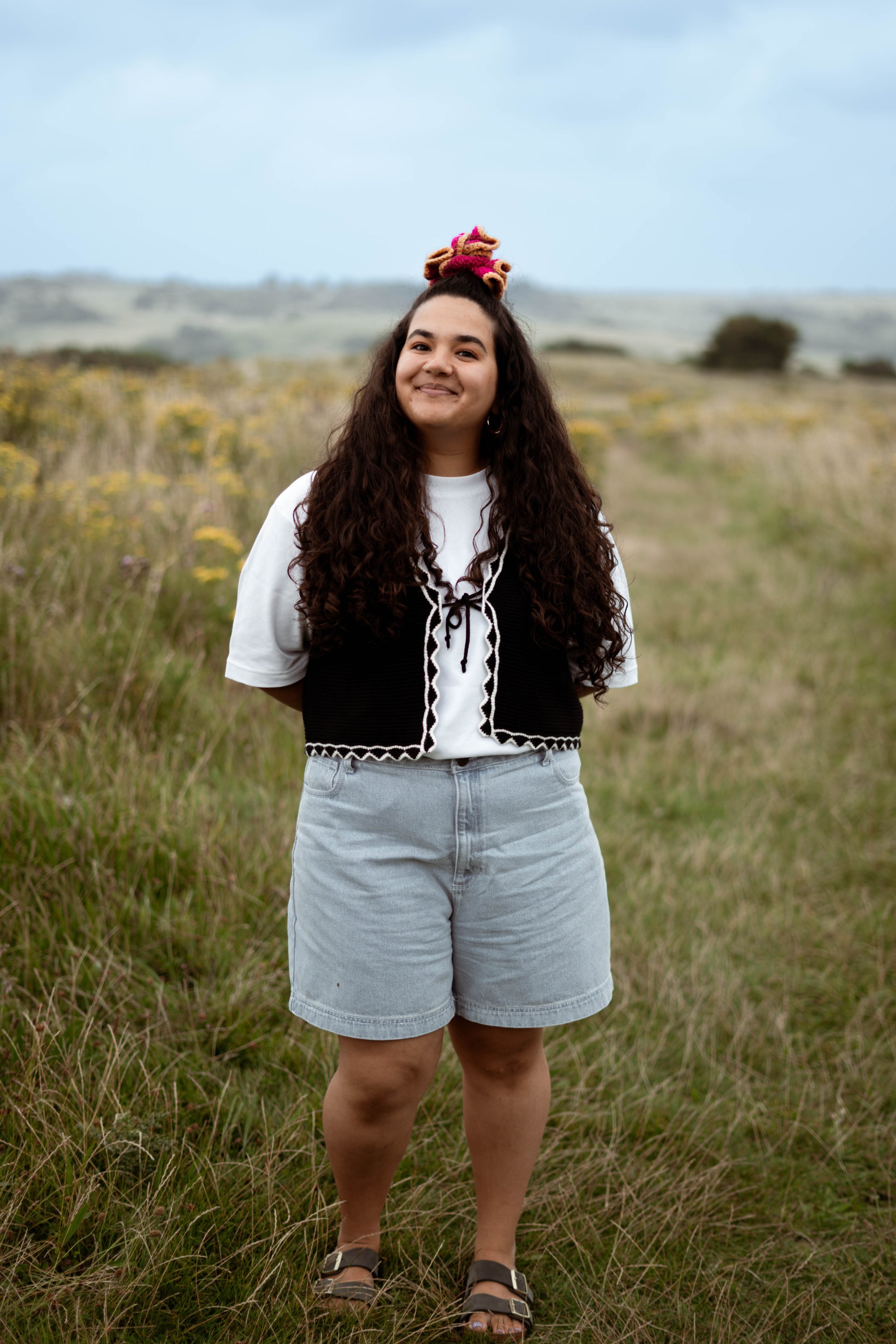 A young woman with long curly hair, wearing a white t-shirt, black vest with white trim, light gray shorts, and sandals, standing outdoors in a grassy field with yellow flowers and rolling hills in the background on a cloudy day.