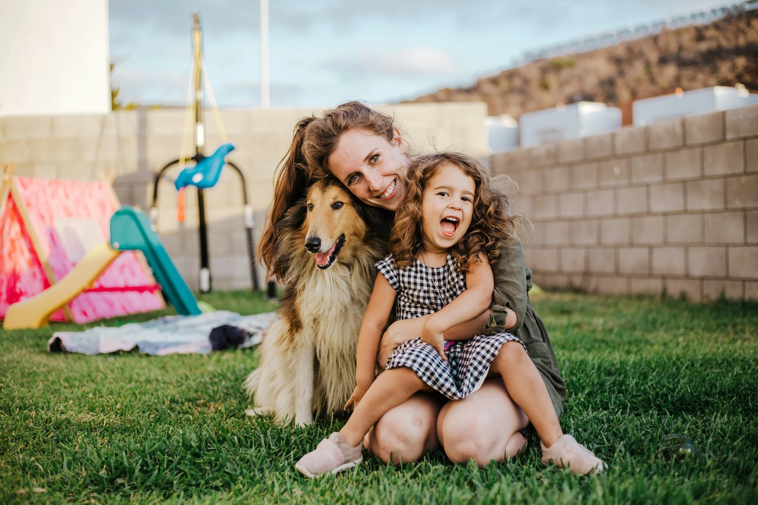 A woman and a young girl sitting on grass in a backyard, hugging a collie dog. The backyard has a brick wall, playground equipment, and a hillside in the background.