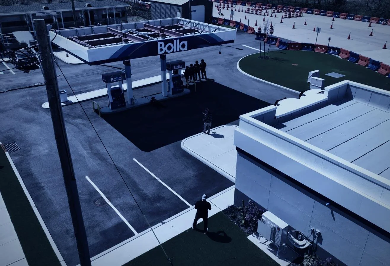 Drone aerial by CXIX: a rooftop scene showing a Bolla gas station with two fuel pumps, a small group of people standing nearby, a person walking on the sidewalk, and a man taking a photo. The area is surrounded by orange traffic barrels and parking s