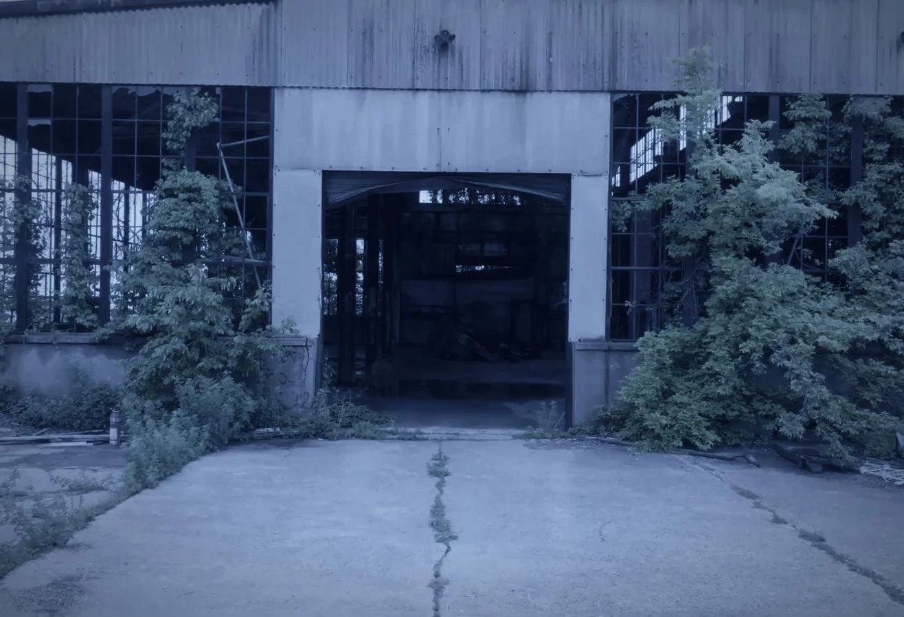 Drone aerial by CXIX: an abandoned building with boarded-up windows, overgrown bushes, and a cracked concrete driveway leading to the entrance.