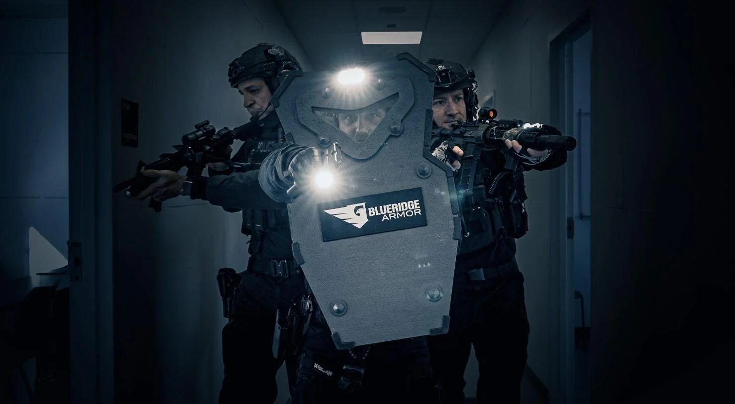 Three police officers in tactical gear, holding rifles and aiming down a hallway, with one officer in front carrying a ballistic shield labeled "BlueRidge Armor."