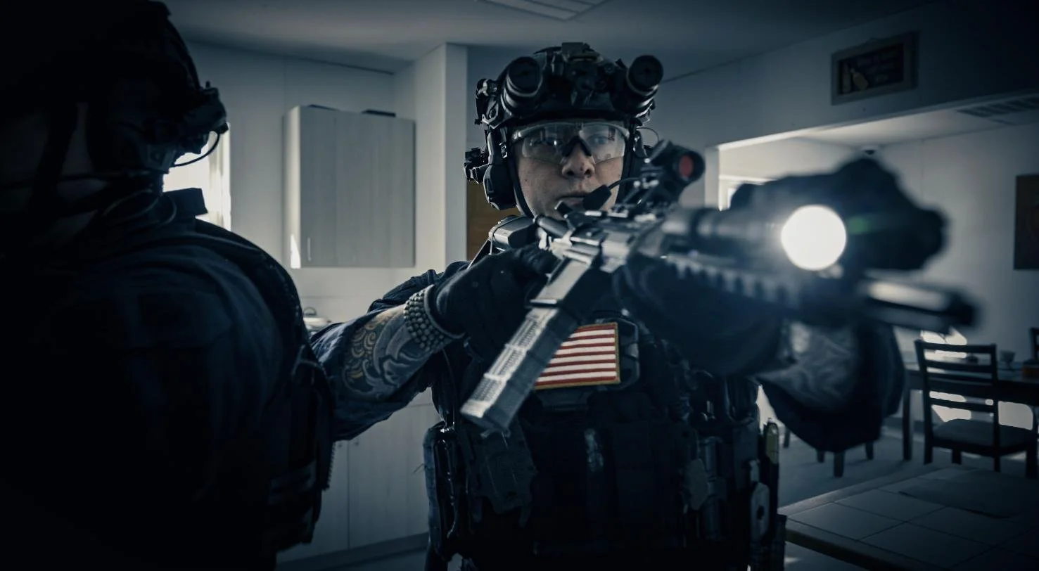 Two tactical police officers in gear, one aiming a firearm with a flashlight attached, inside a room with chairs and cabinets.