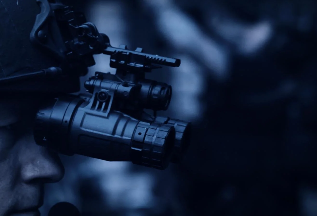 Nocturn Industries night vision shoot by CXIX: close-up of a person wearing night vision goggles with a mounted scope, looking through them in a dark setting.