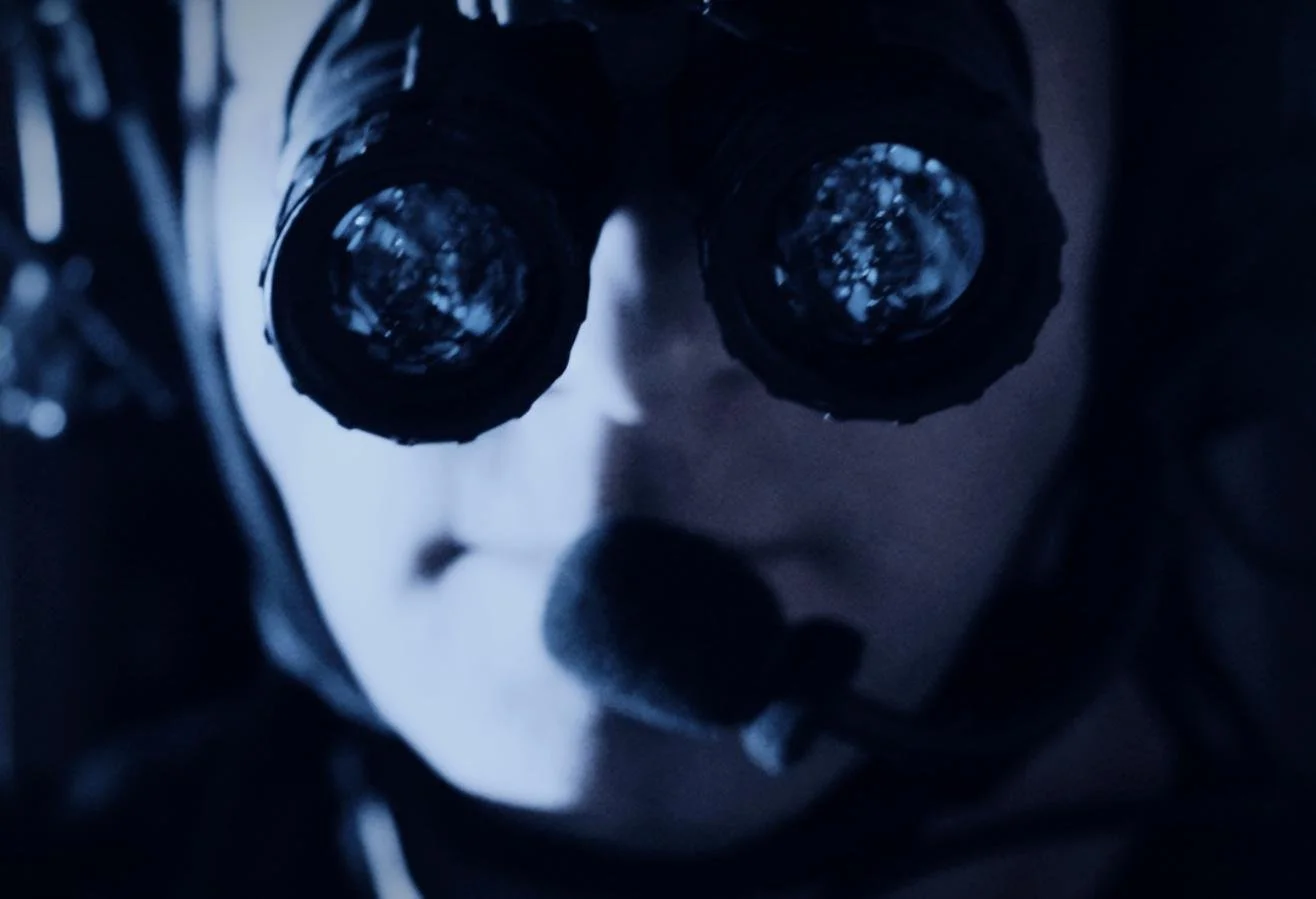 Nocturn Industries night vision shoot by CXIX: close-up of a doll's face with binoculars covering its eyes, in a dark setting with bluish lighting.