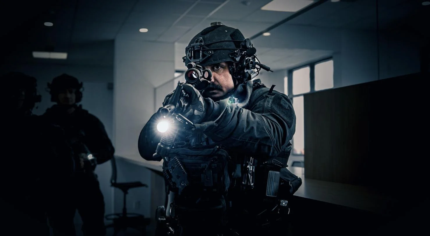 Long Island SWAT Challenge, shot by CXIX: a soldier in tactical gear aiming a firearm with a flashlight attached while wearing a helmet with night vision goggles, inside a modern building with windows in the background.