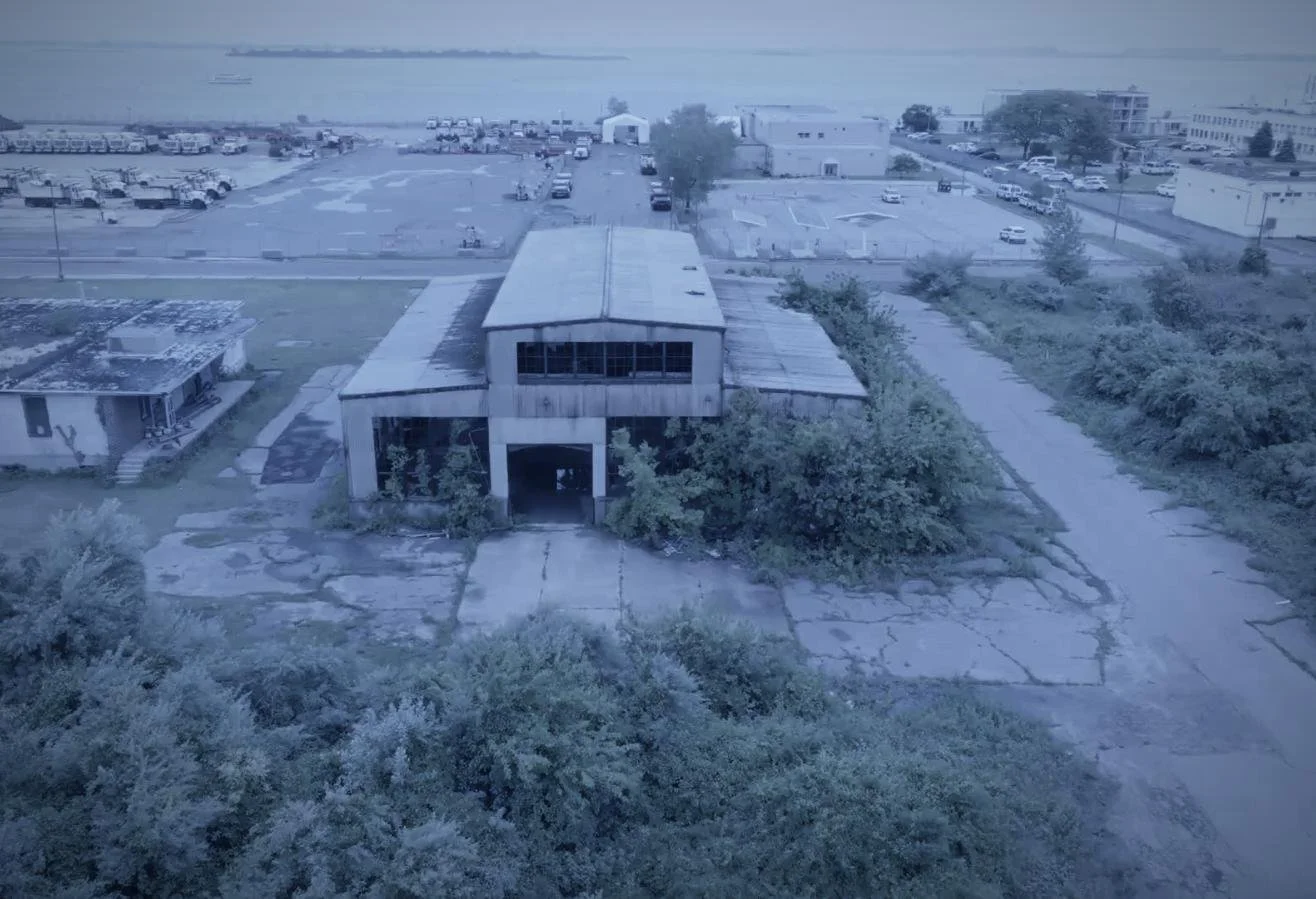 Drone aerial by CXIX: an abandoned, weathered building with overgrown bushes and cracked concrete in front, surrounded by parking lots and other structures, near a body of water.