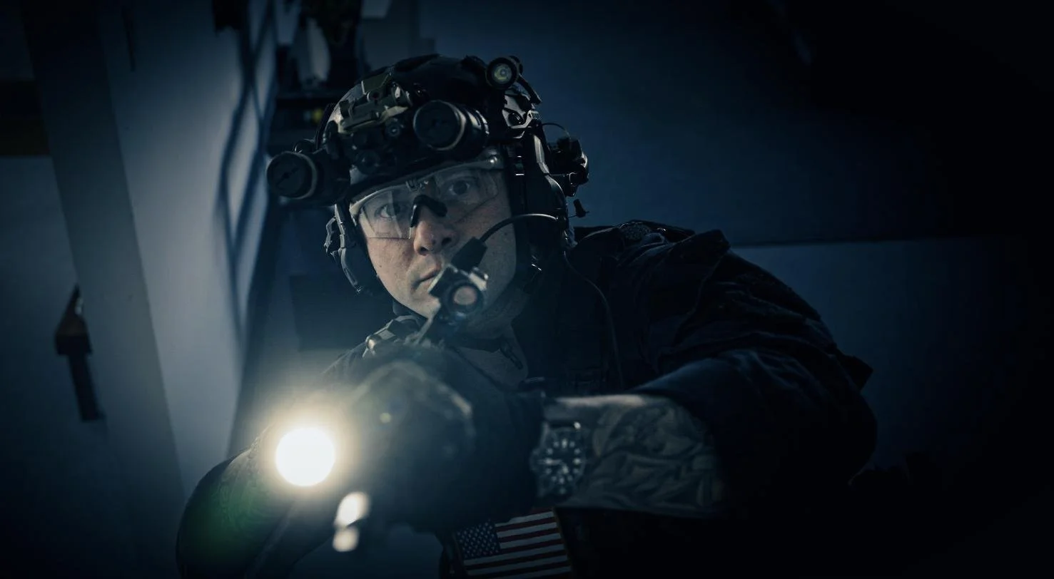 A soldier wearing night vision gear and camouflage, holding a flashlight and aiming a weapon in a dark environment.
