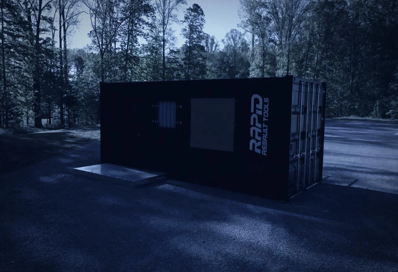 Drone aerial by CXIX: a black shipping container labeled 'RASP Assault Tools' placed on a paved area outdoors with trees and a clear sky in the background.