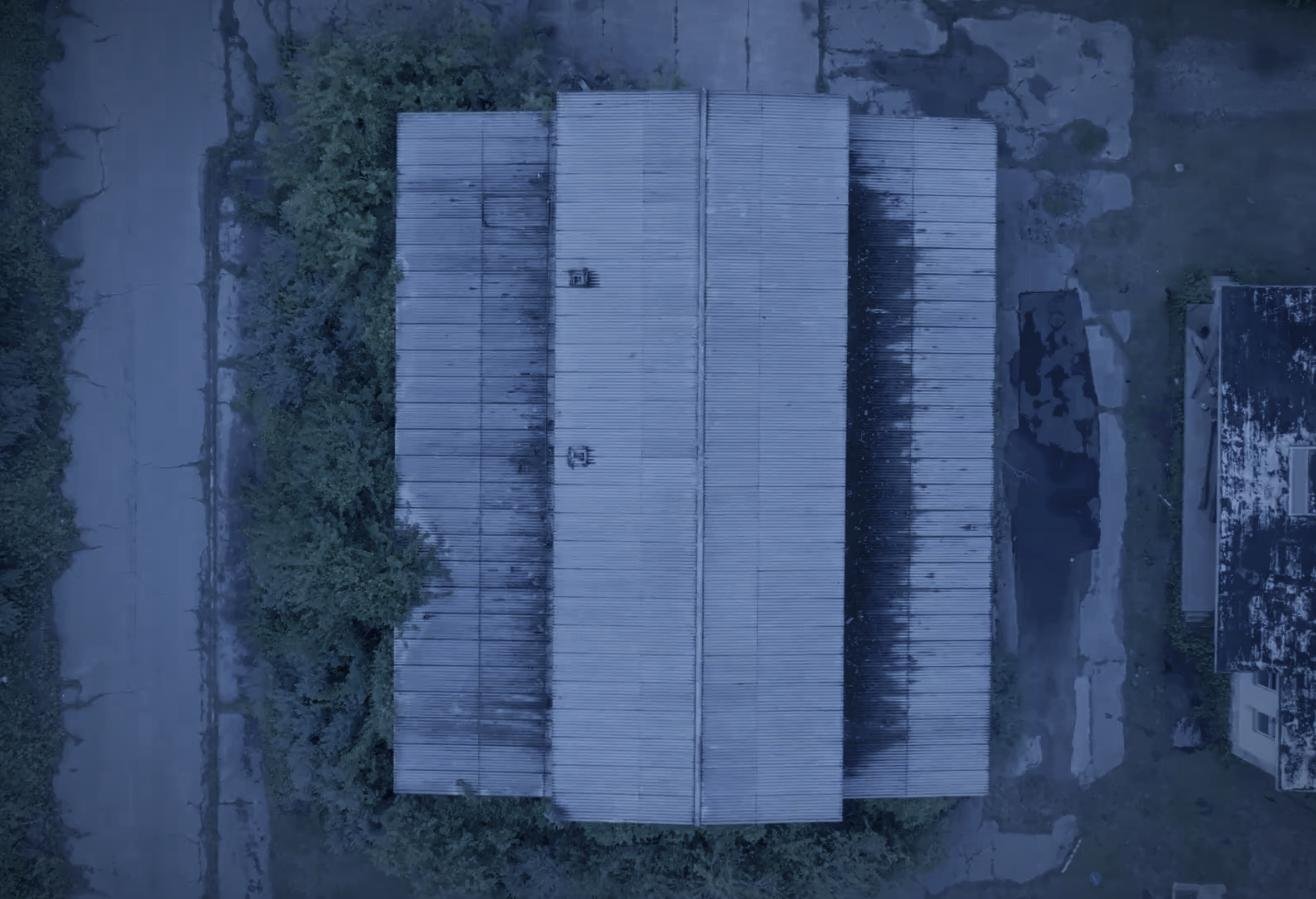 Drone aerial by CXIX: top-down view of an old, weathered metal building with a corrugated roof, surrounded by trees and adjacent to a stone or concrete structure.
