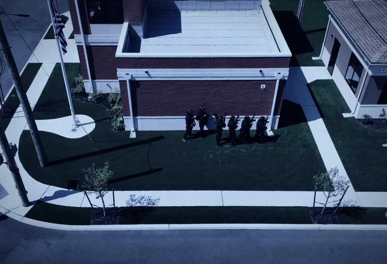 Drone aerial by CXIX: an aerial view of a building with a group of uniformed police officers walking in a line on the sidewalk outside.