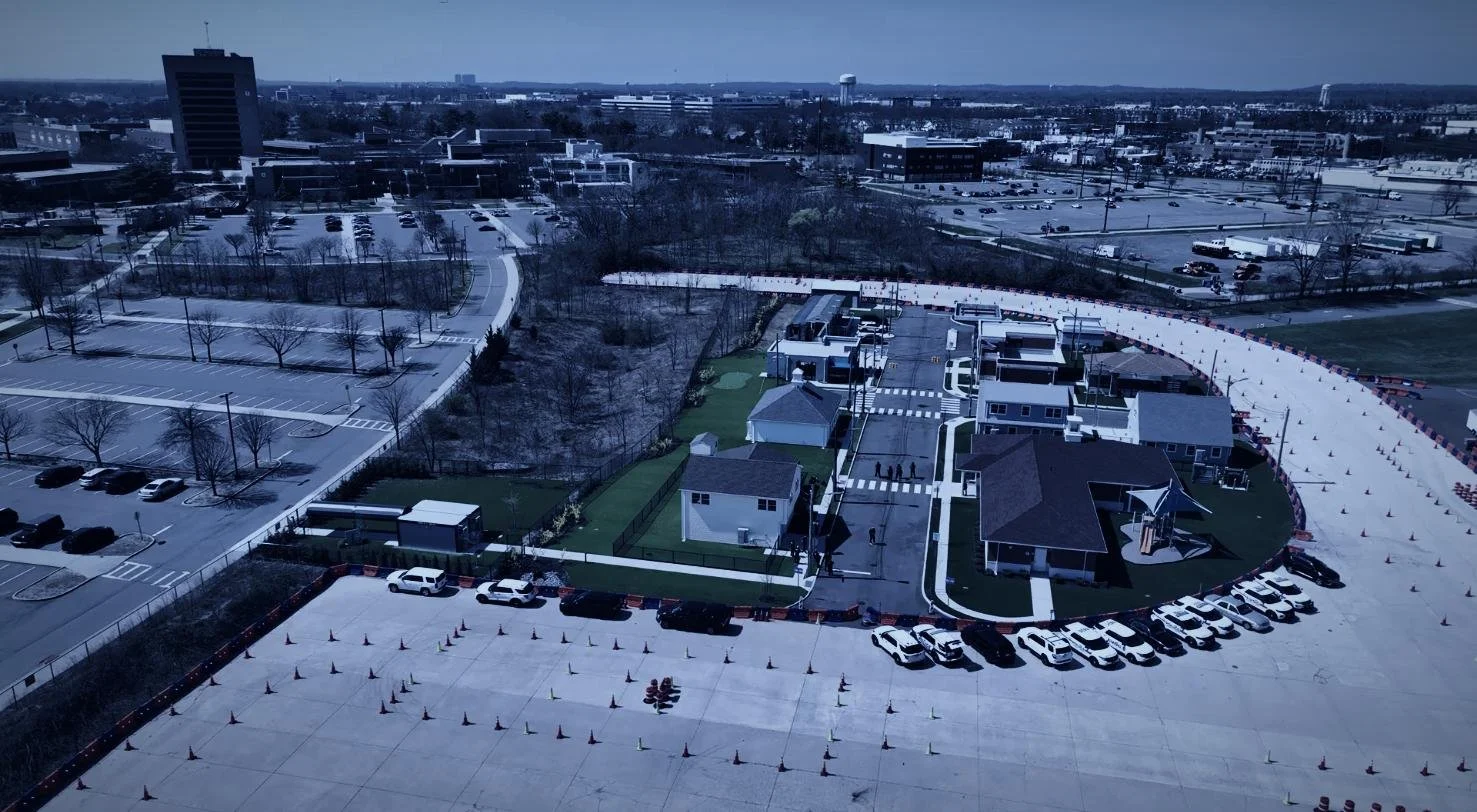 Long Island SWAT Challenge, shot by CXIX: aerial view of a commercial and parking lot area with some residential structures, trees, and a large empty parking lot with traffic cones, under a cloudy sky.