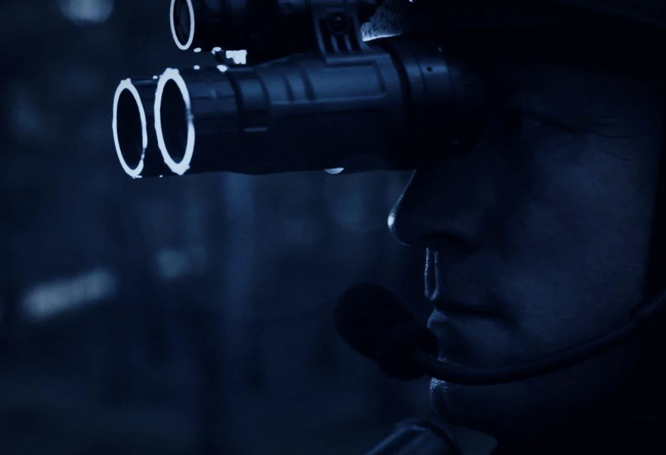 Nocturn Industries night vision shoot by CXIX: a person looking through a telescope or binoculars in a dark environment, illuminated by minimal blue lighting.