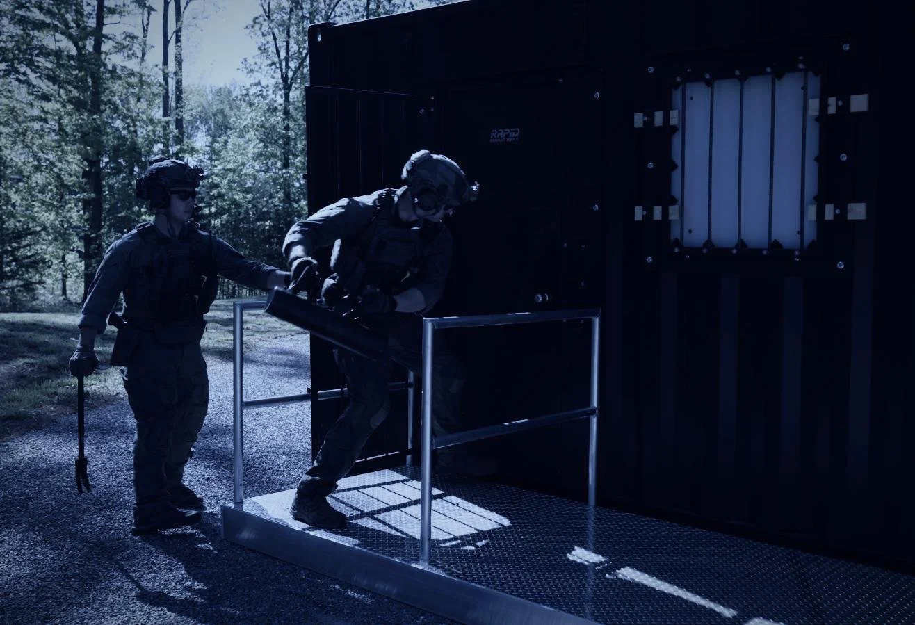 Rapid Assault Tools shoot by CXIX: two armed military personnel in tactical gear, one assisting the other as they step onto a platform in front of a black container or vault outdoors, surrounded by trees.