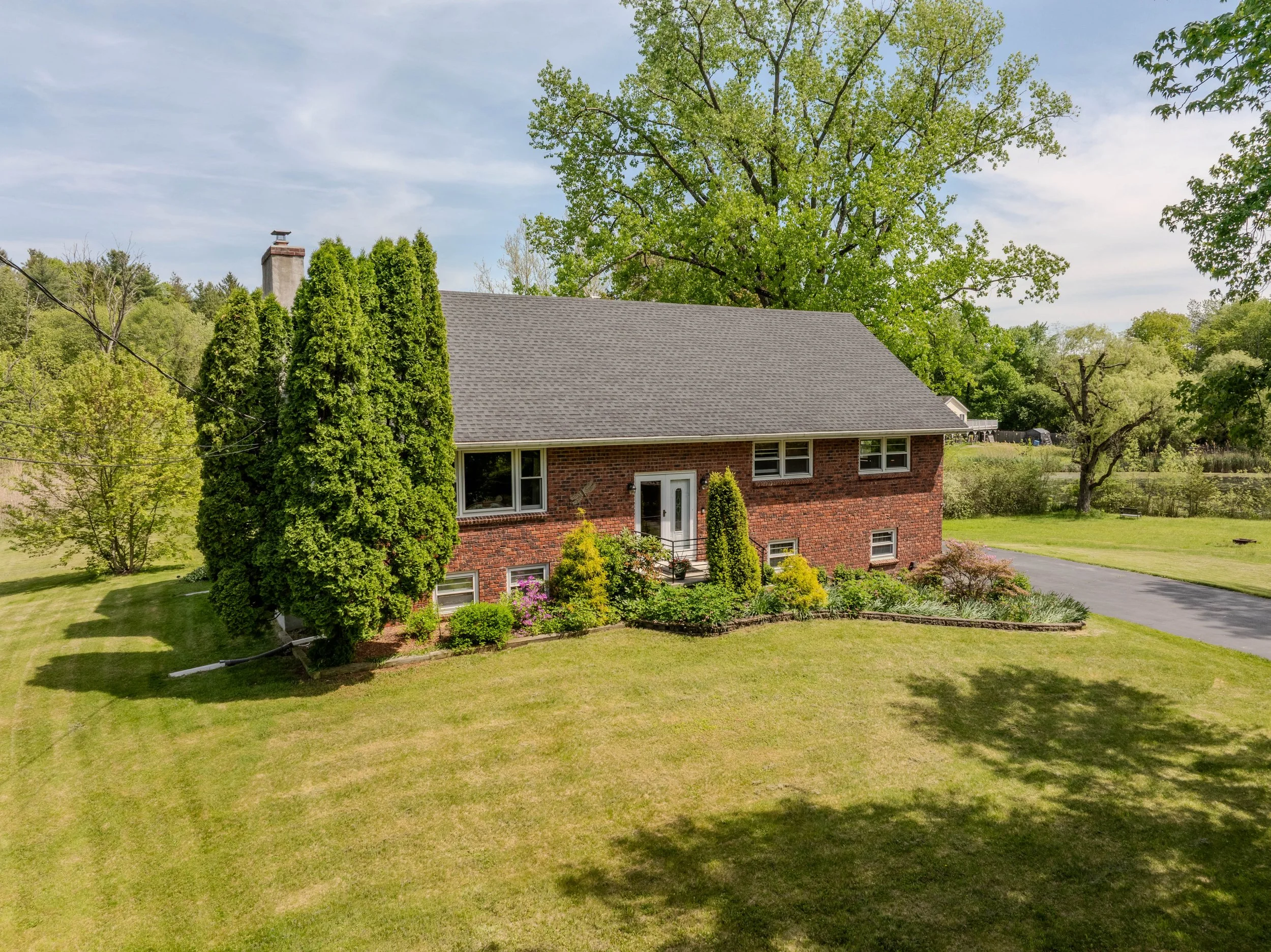 A two-story brick house with a gray shingled roof, surrounded by a lush green yard with trees and shrubbery, under a partly cloudy sky.