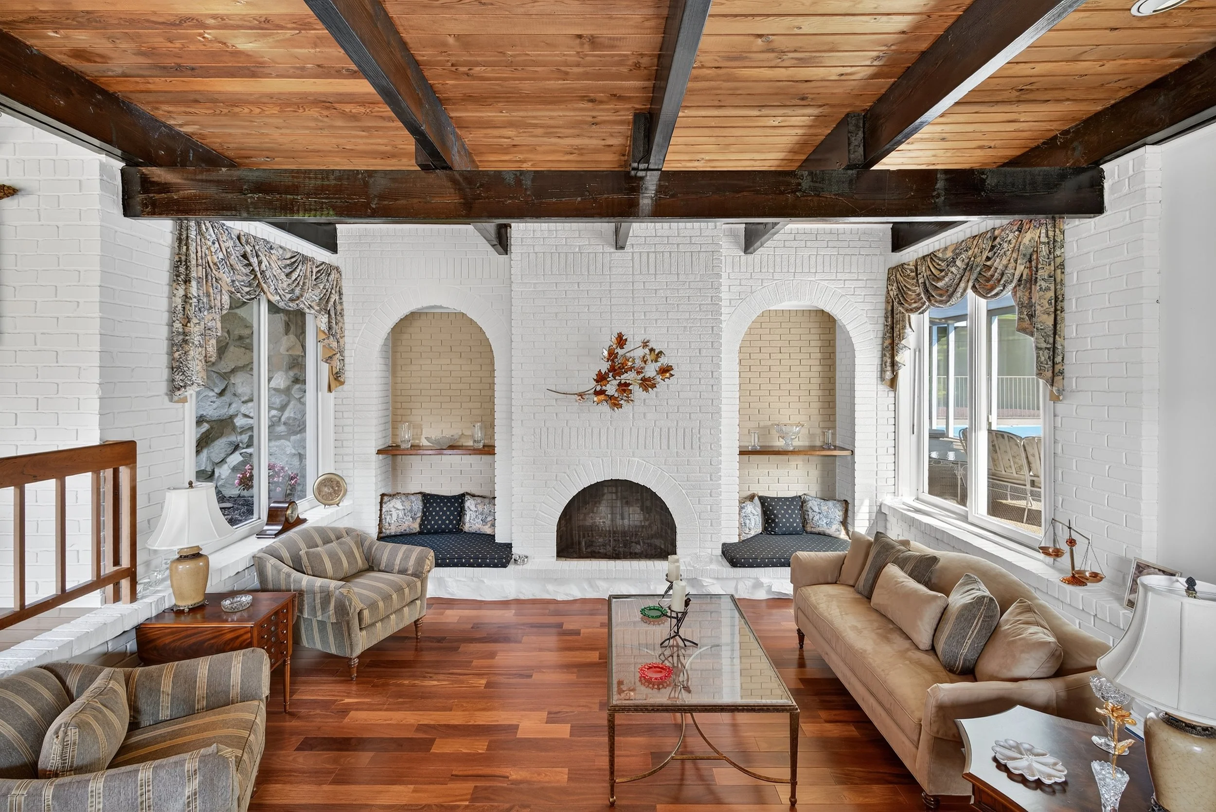Living room with white brick fireplace, built-in window seats, floral curtains, wooden ceiling with exposed beams, hardwood floors, sofas, armchair, coffee table, and decorative items.