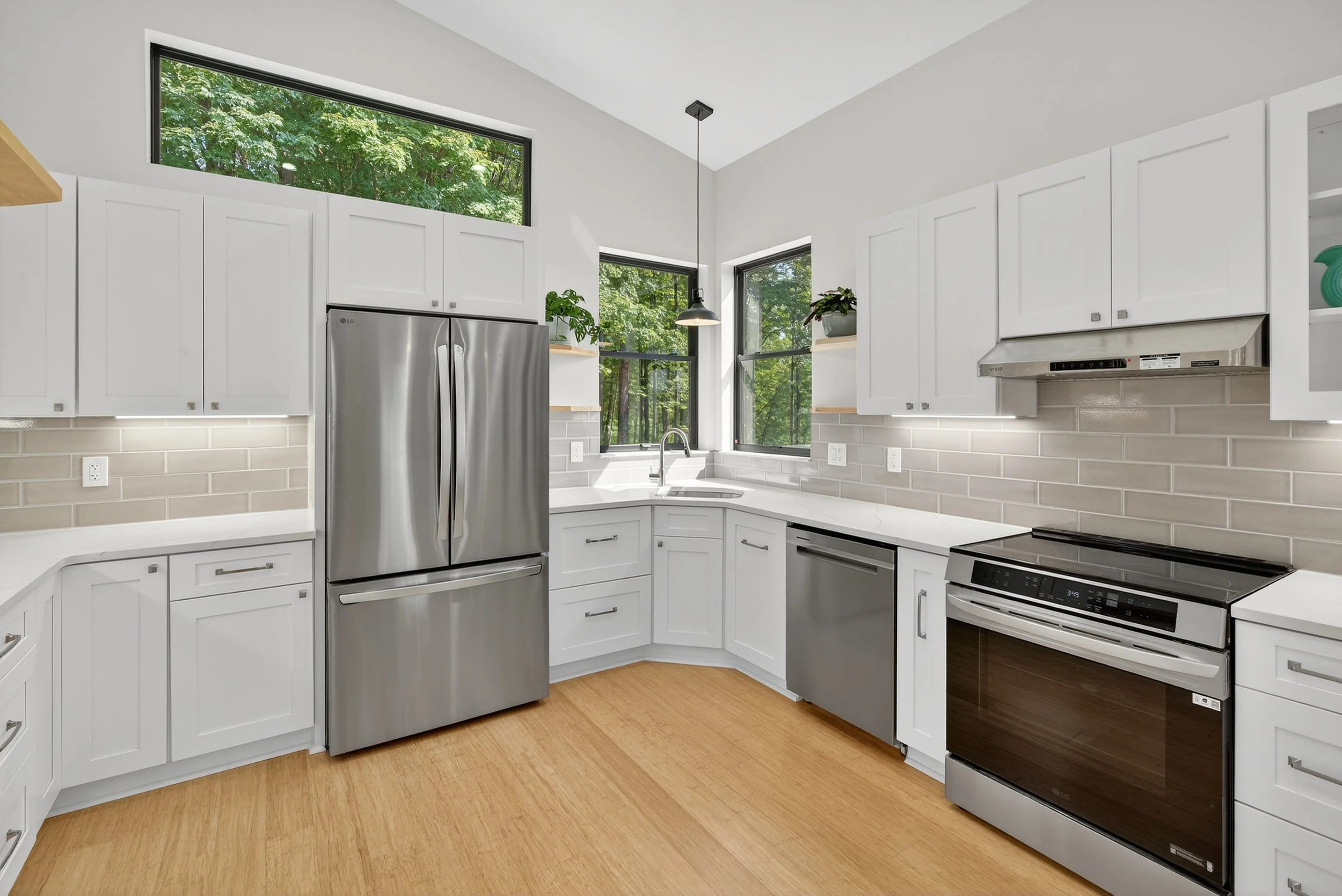 Modern kitchen with white cabinets, stainless steel refrigerator, oven, and dishwasher, surrounded by large windows showing green trees, light wood flooring, and minimal decor.