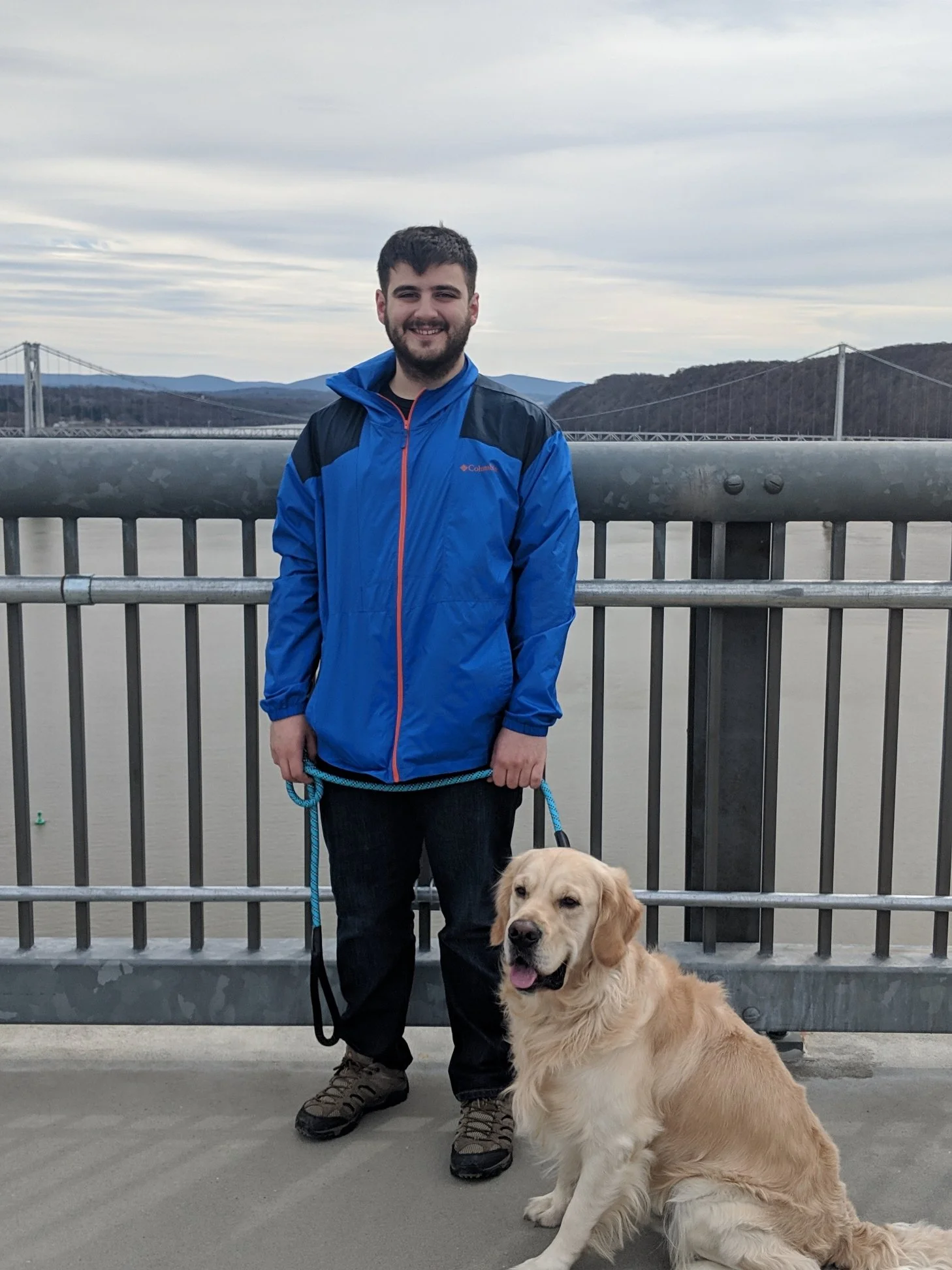 Man in blue jacket holding a leash with a sitting golden retriever on a bridge over a body of water, with hills and a suspension bridge in the background.