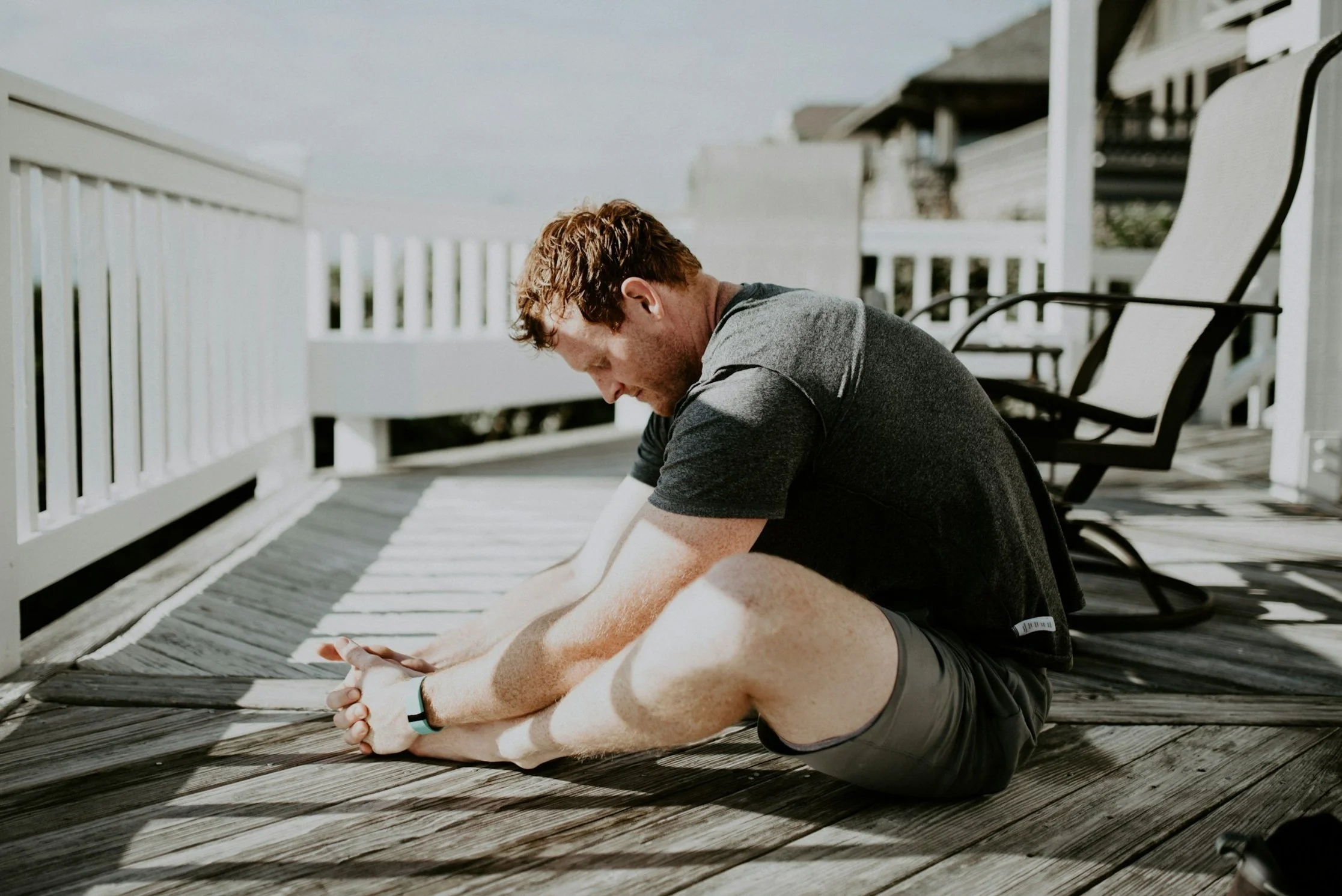 Man sitting on a wooden deck, stretching his legs while leaning forward with hands clasped, under bright daylight.