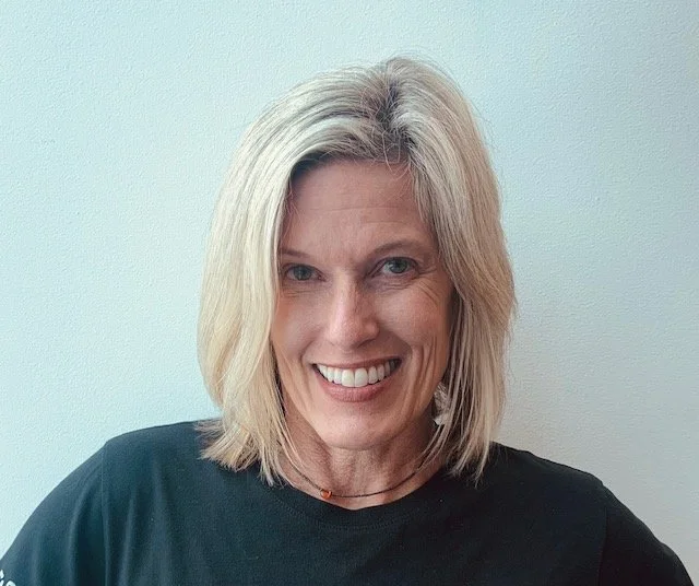 A woman with shoulder-length blonde hair, smiling, wearing a black shirt, standing against a plain light-colored wall.