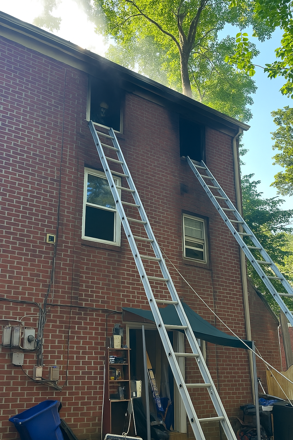 The back of a brick multi-story house with two extension ladders leaning against the wall, reaching the upper windows, and a firefighter peering out of a smoke-damaged second-story window, suggesting a recent or ongoing fire emergency.