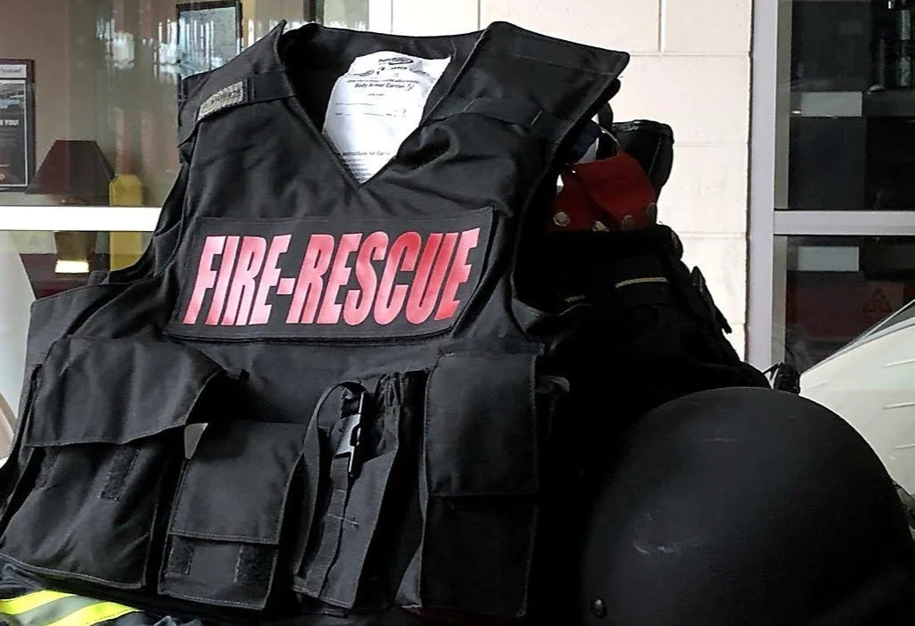 A black firefighter's vest with a pink and white 'FIRE-RESCUE' patch, placed on a surface with a motorcycle helmet nearby, in front of a building with glass and brick walls.