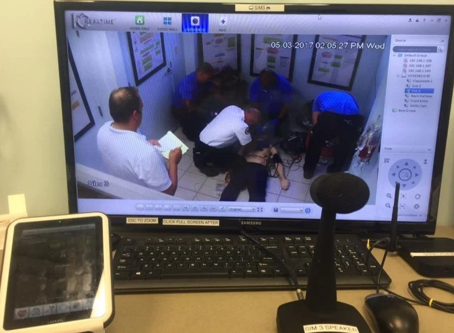 Surveillance footage showing five people in a small room with posters on the wall. One person appears to be unconscious or injured on the floor, while others attend to them, with one person holding a clipboard.