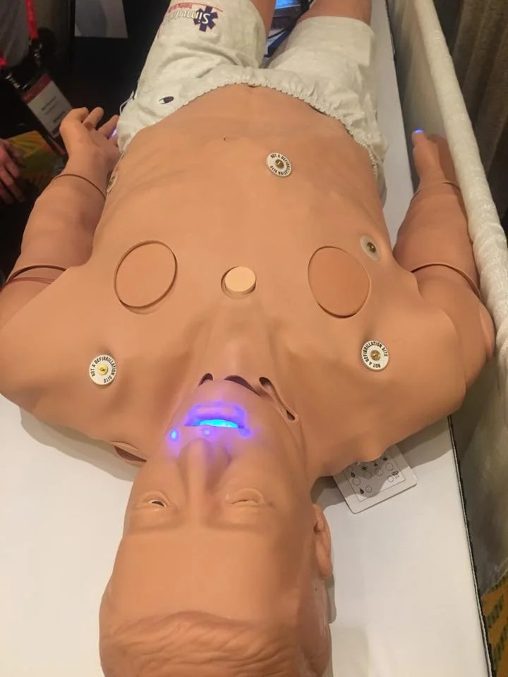 Medical training mannequin lying on a bed with sensors and a glowing blue mouth, used for practicing CPR and emergency procedures.