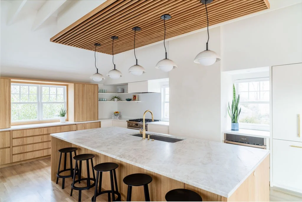 Wide view of modern kitchen with white oak island, quartzite countertop, and slatted oak ceiling feature.