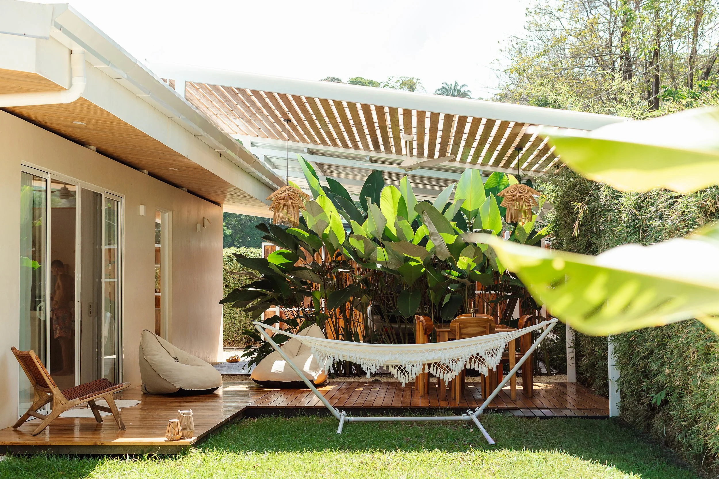 Patio con zona de descanso, hamaca blanca, decoración con plantas grandes, mesas de madera, sombrillas de paja y mobiliario de madera, en un entorno exterior con vegetación pavones costa rica surf