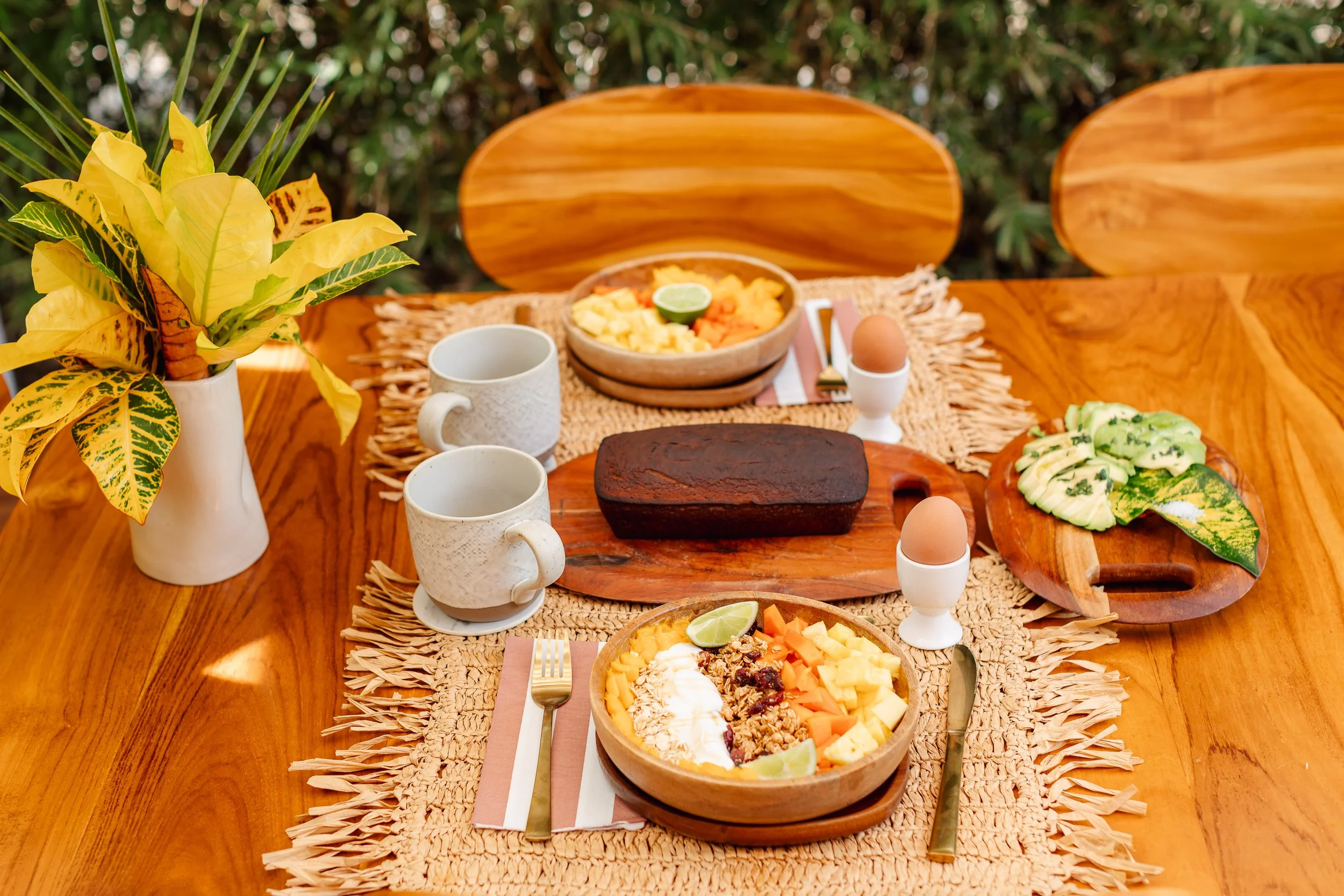 Mesa de desayuno con tazas, yogur con frutas, panqueques con frutas, huevo cocido, un pastel y una planta