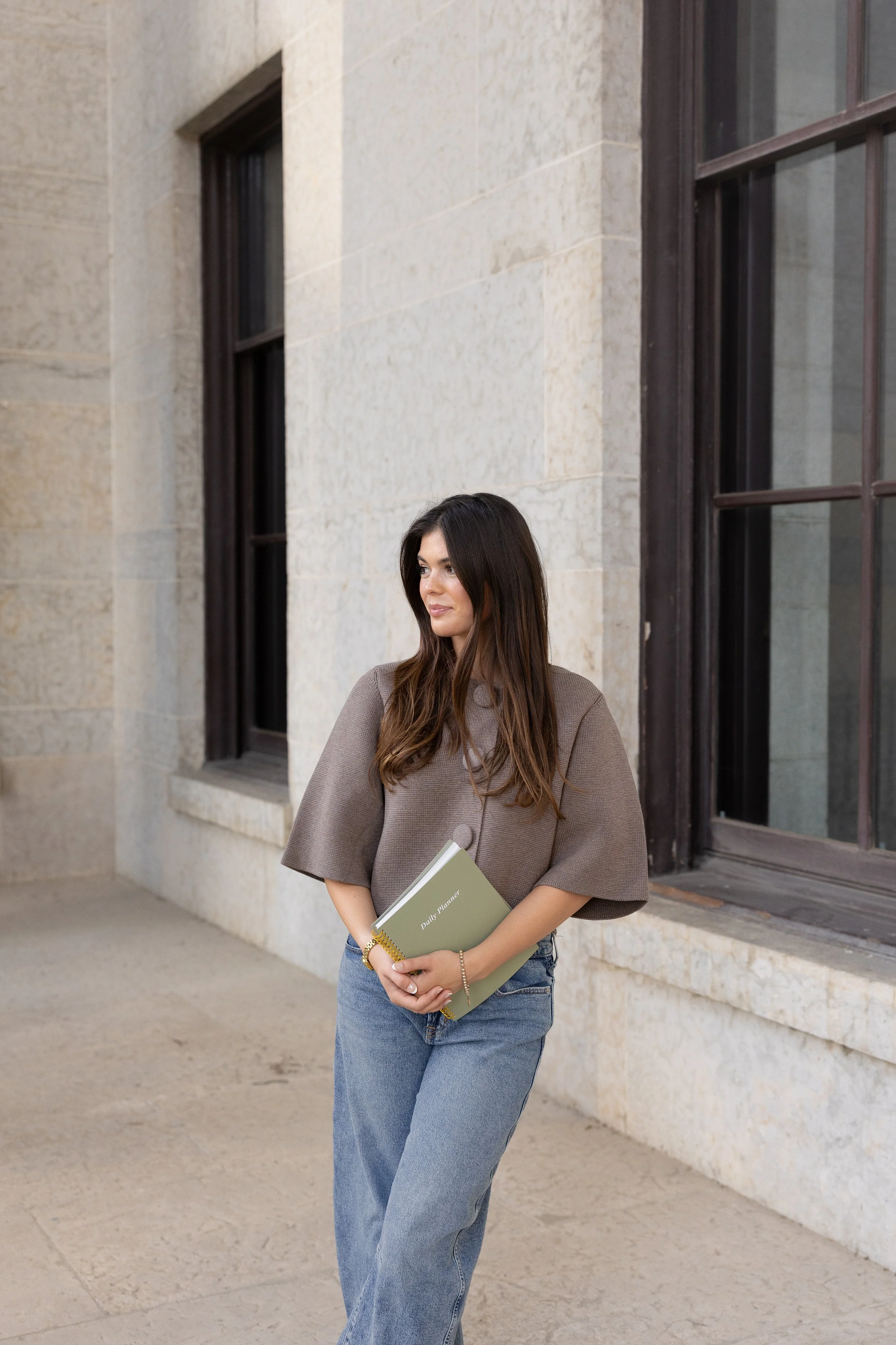 A woman stands outside a building with stone walls and large windows, holding a green textbook titled 'Beauty Planner' and looking to the side.