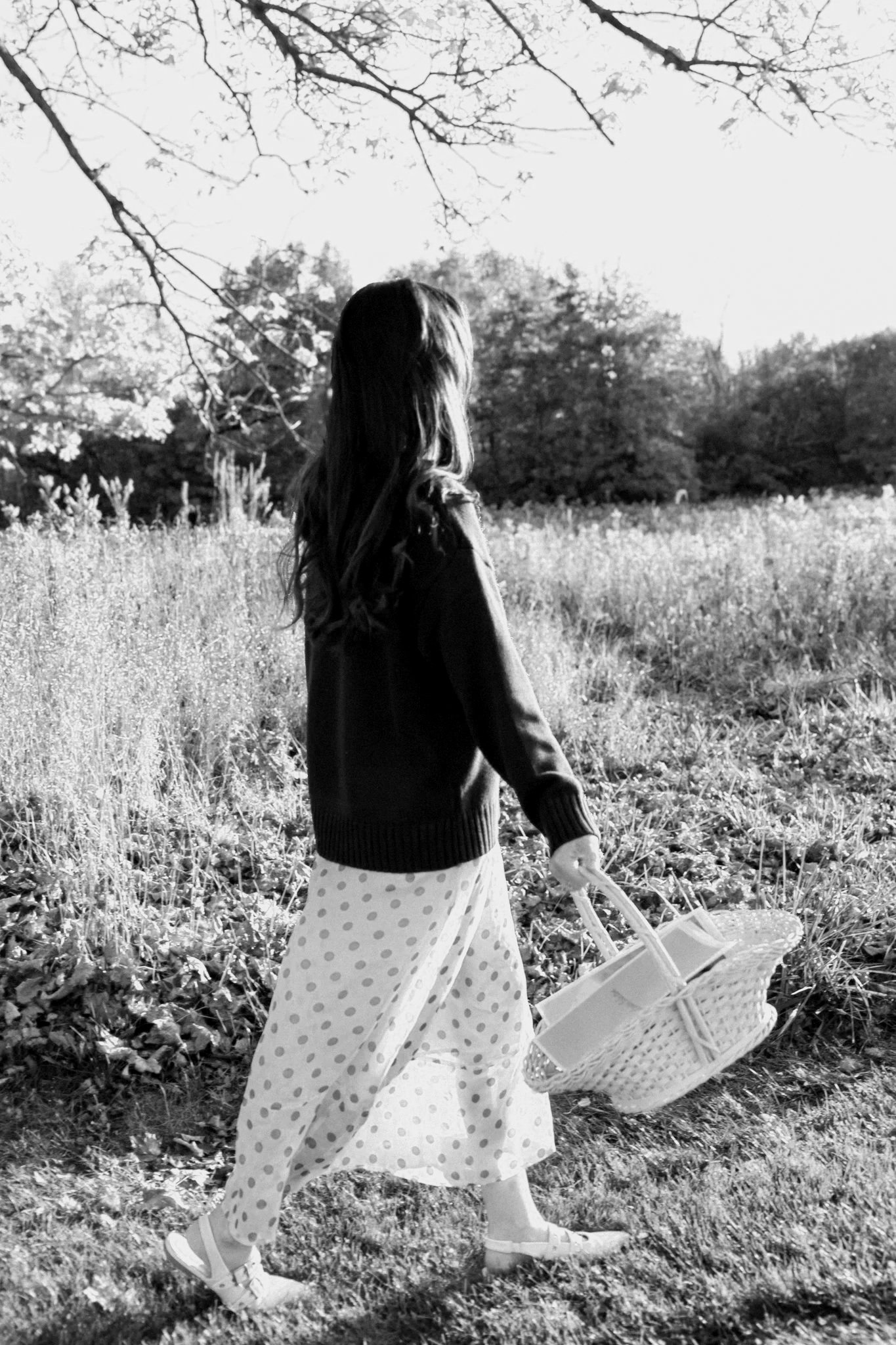 Black-and-white photo of a woman in a polka-dot dress carrying a wicker basket through a sunny meadow.