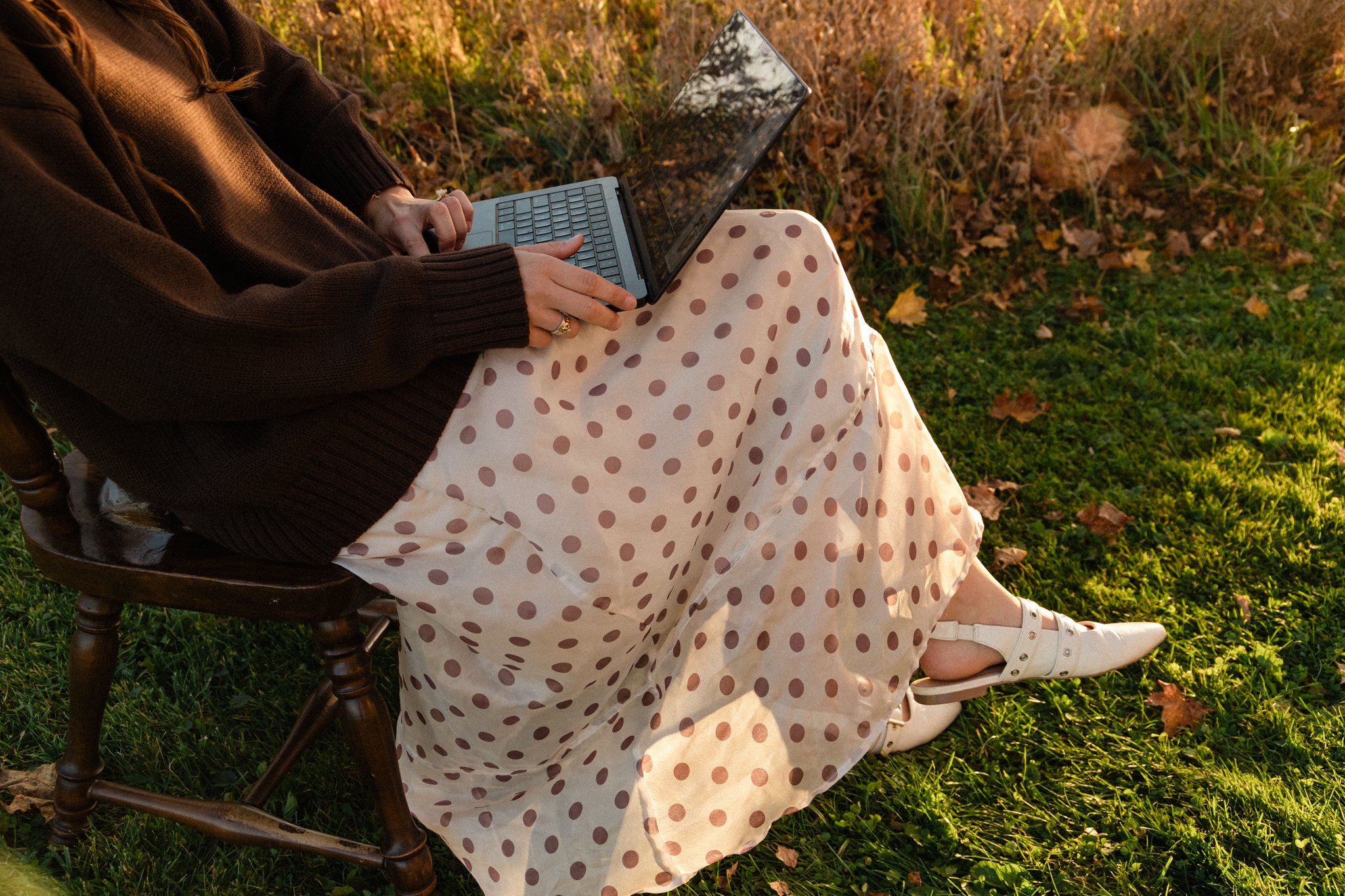 Woman using a laptop outdoors while seated on a wooden chair in a sunlit grassy yard
