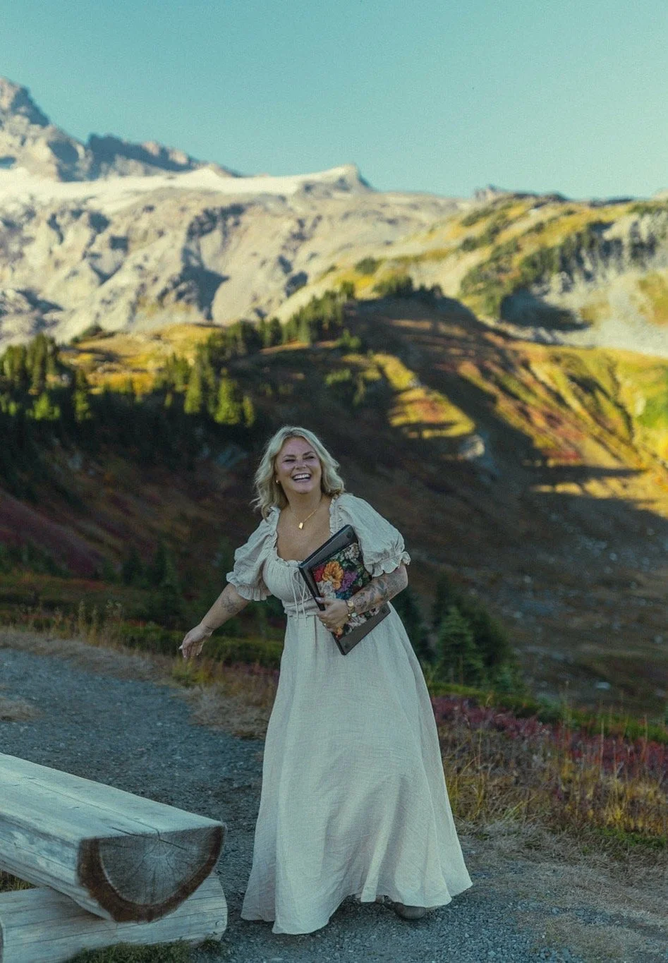 Smiling woman in a long white dress holding a floral book outdoors with rugged mountain peaks and alpine trees behind her