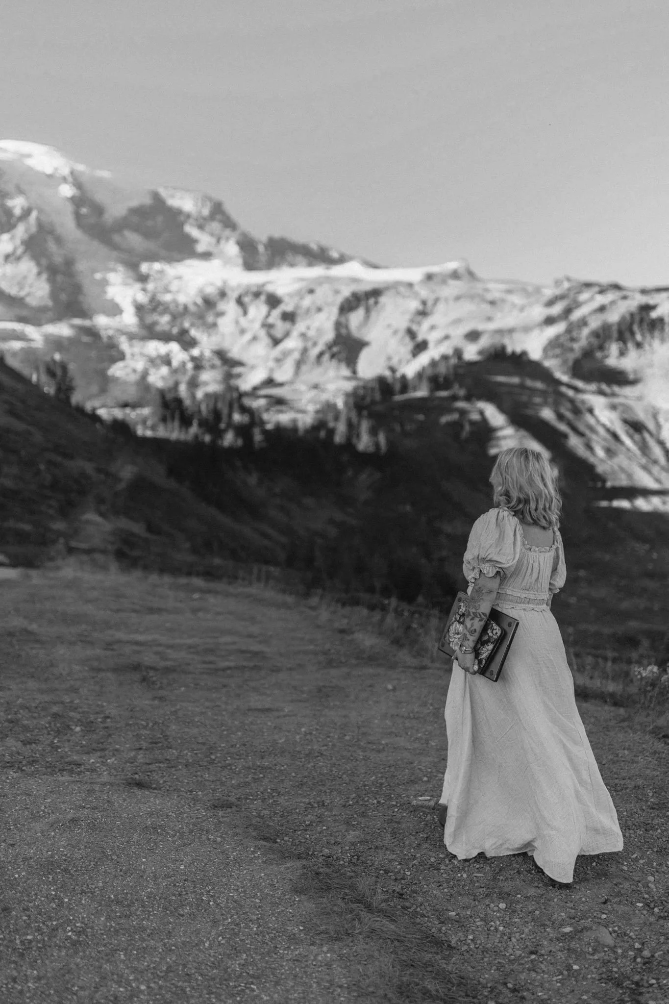 Woman in a long dress holding framed artwork on a rocky landscape overlooking snow-covered mountains