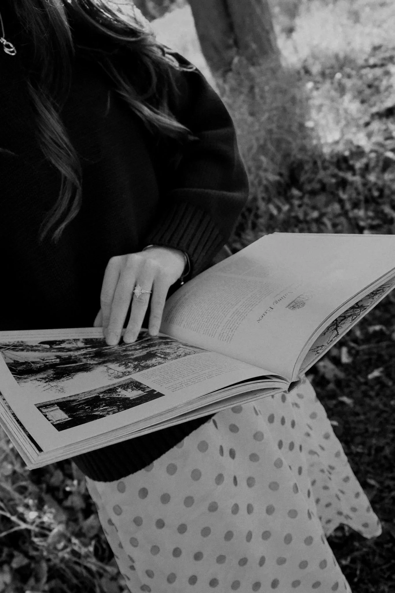 Black-and-white photo of a woman reading an open illustrated book outdoors.