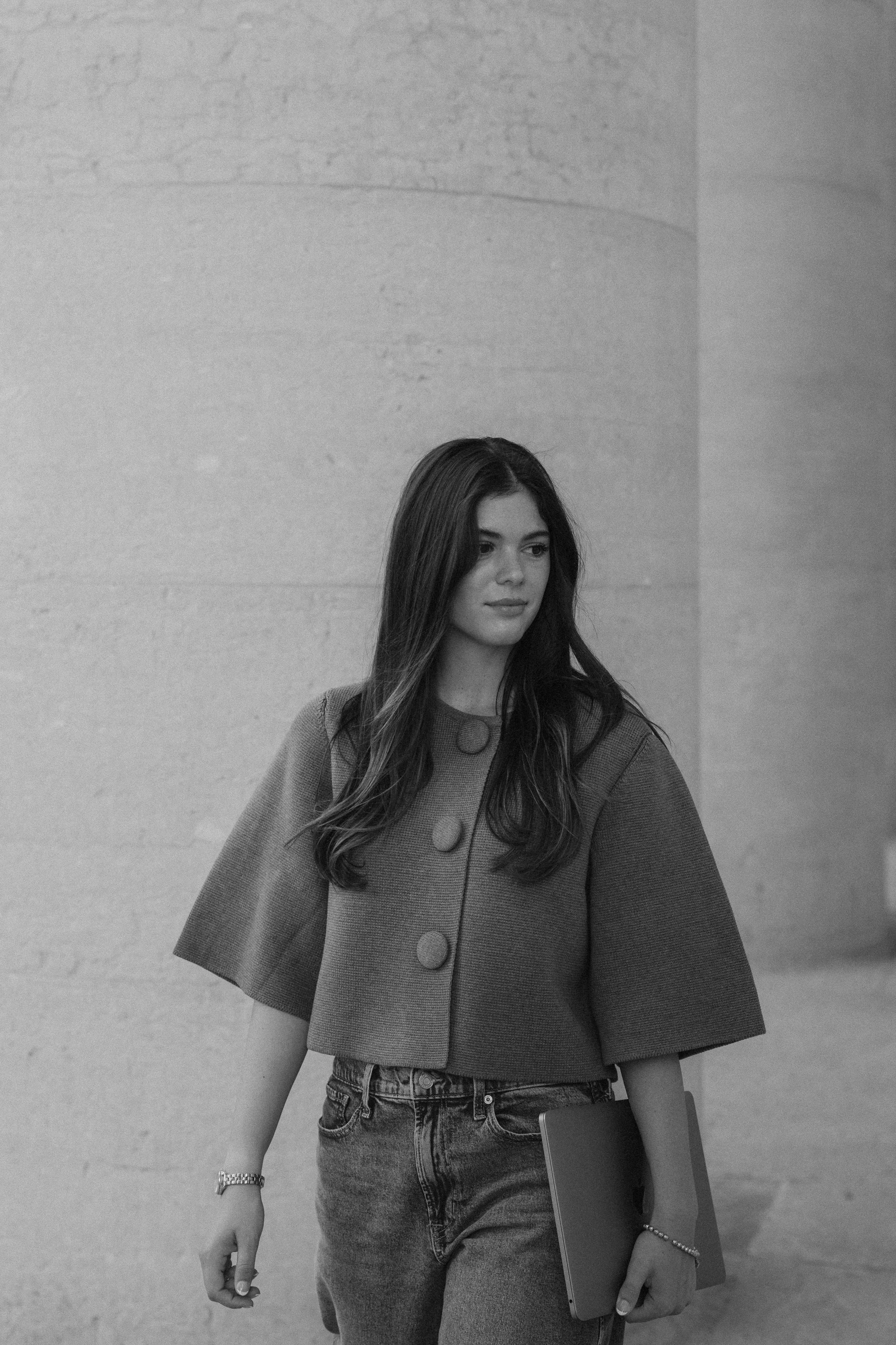 A young woman with long dark hair wearing a short-sleeved, buttoned jacket and jeans, holding a laptop and walking indoors.