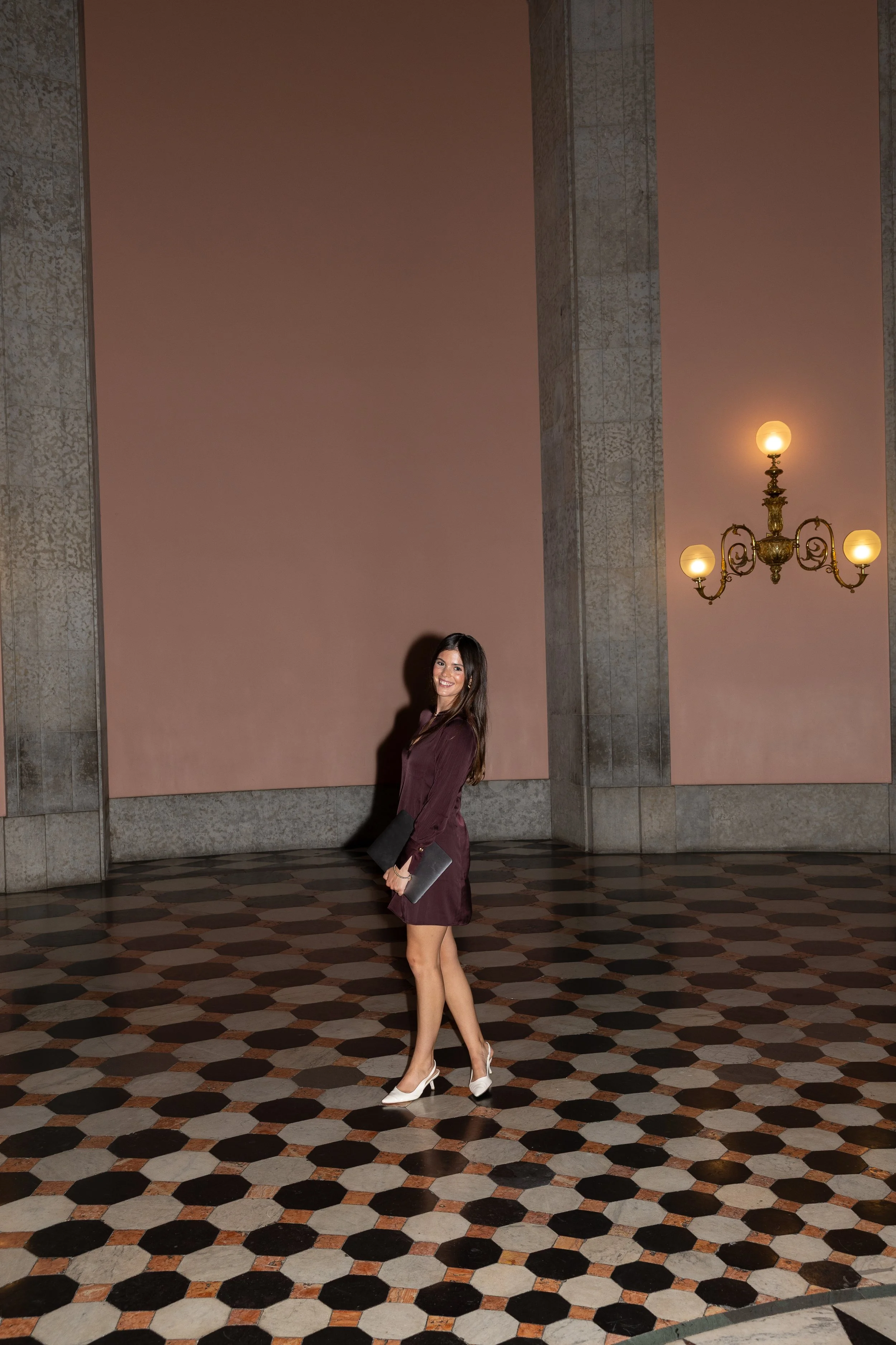A woman in a burgundy dress and white heels stands smiling in an elegant interior with a patterned tile floor, large pink wall panels, and ornate brass chandeliers.