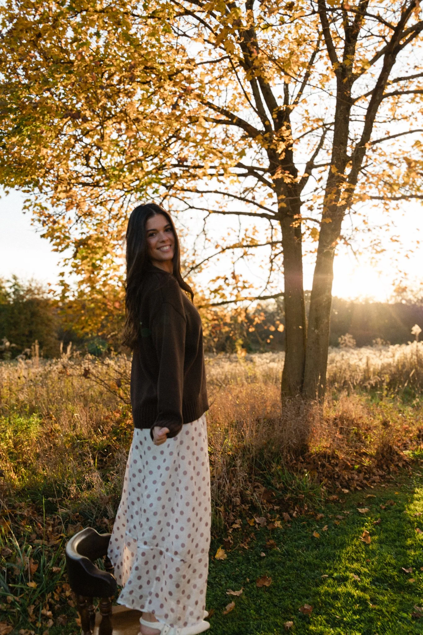 Woman in a dark sweater and polka-dot skirt standing outdoors beneath autumn trees at sunset