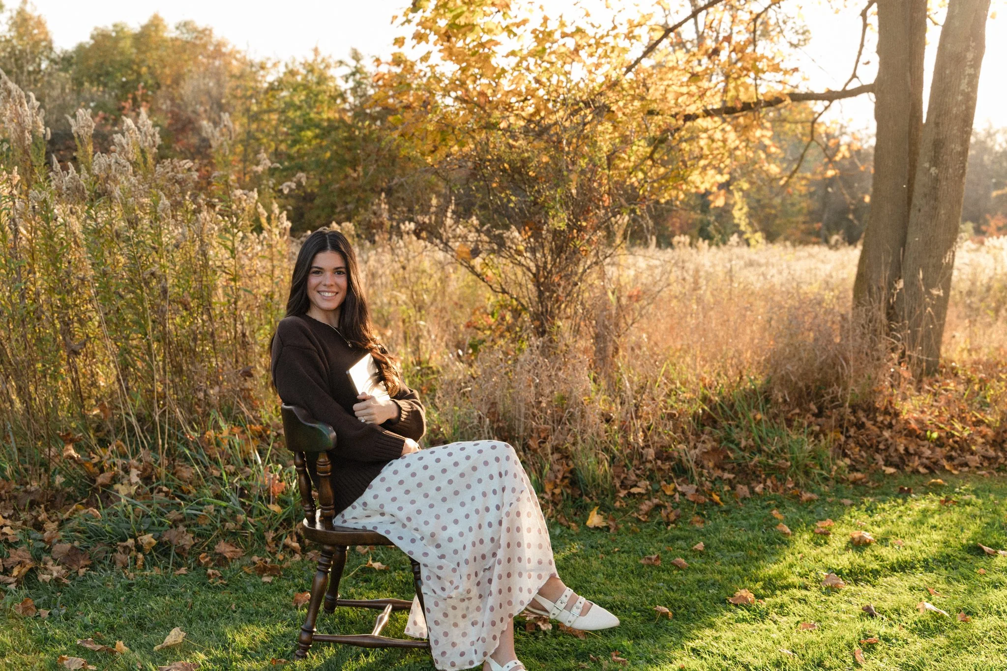 Smiling woman sitting in a wooden rocking chair outdoors in a sunlit autumn meadow, wearing a black sweater and polka-dot skirt.