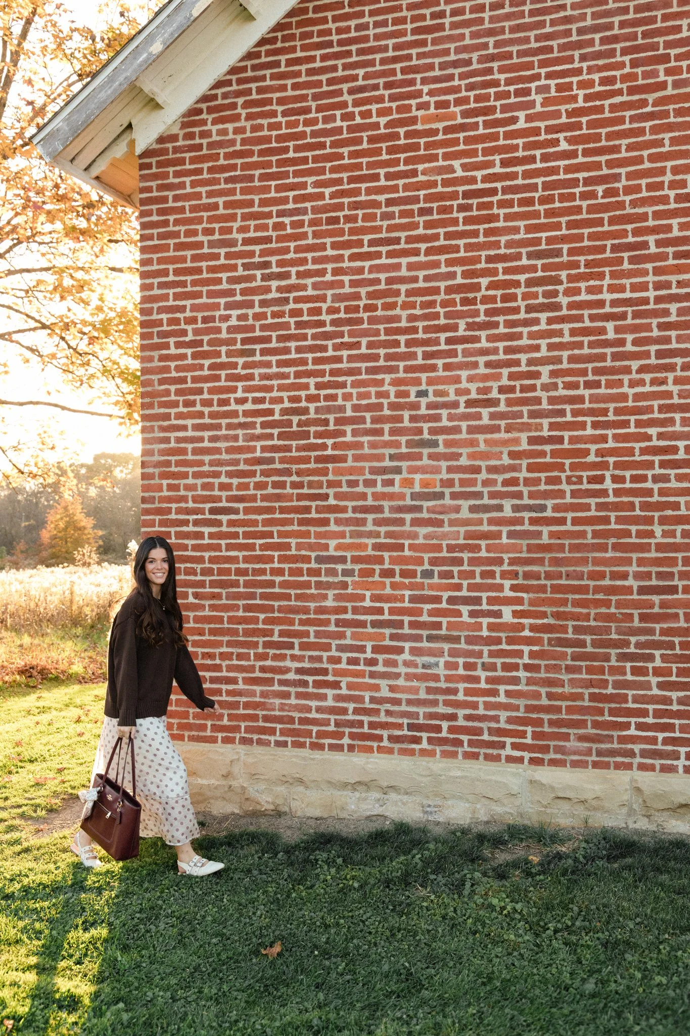 Smiling woman in a black sweater and polka-dot skirt standing beside a red brick building in autumn sunlight
