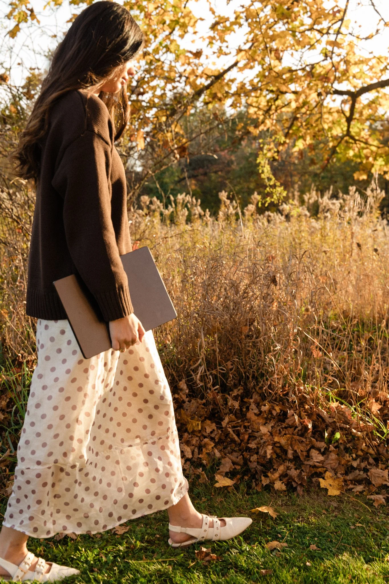 Woman carrying a black book through an autumn woodland, wearing a brown sweater, polka-dot skirt, and white sandals.