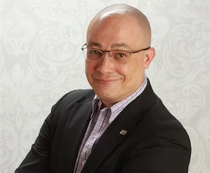 Portrait of a bald man with glasses wearing a dark suit and light checkered shirt, smiling, against a patterned white background.