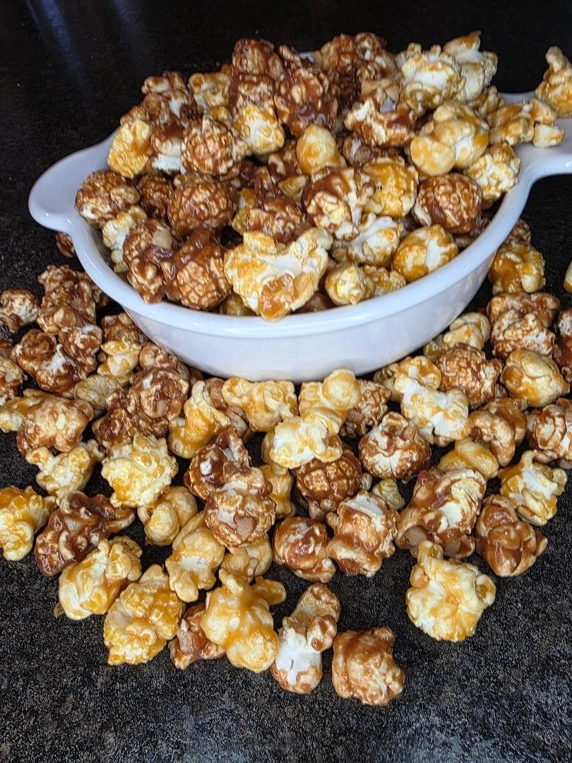Buttered caramel popcorn in a white serving bowl, with some spilled around it on a black surface.